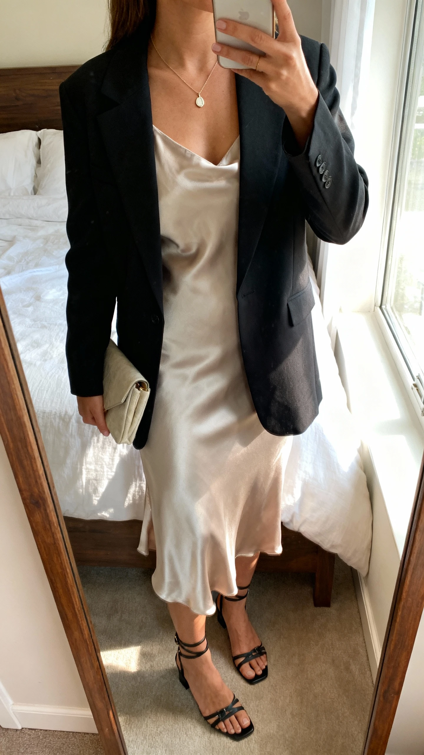 iPhone mirror selfie of a woman wearing a silk slip dress with a tailored blazer layered over it, delicate pendant necklace, small clutch, and strappy heels, face not visible, cropped at neck, bedroom mirror, soft morning window light, iPhone photo quality.