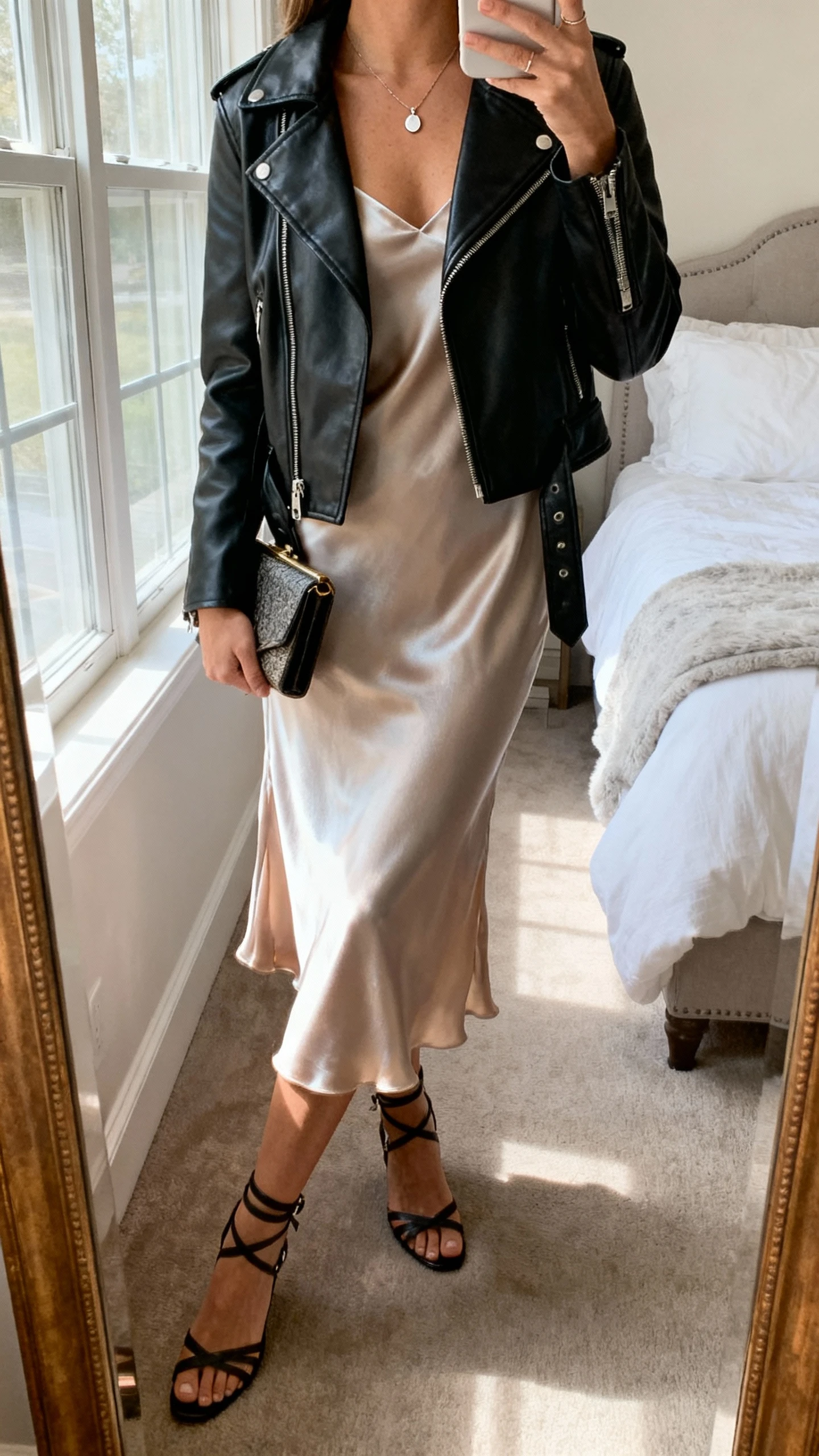 iPhone mirror selfie of a woman wearing a silky slip dress with a black leather moto jacket, strappy heels, small clutch, and delicate pendant necklace, face not visible, cropped at neck, bedroom mirror, natural window light, iPhone photo quality.