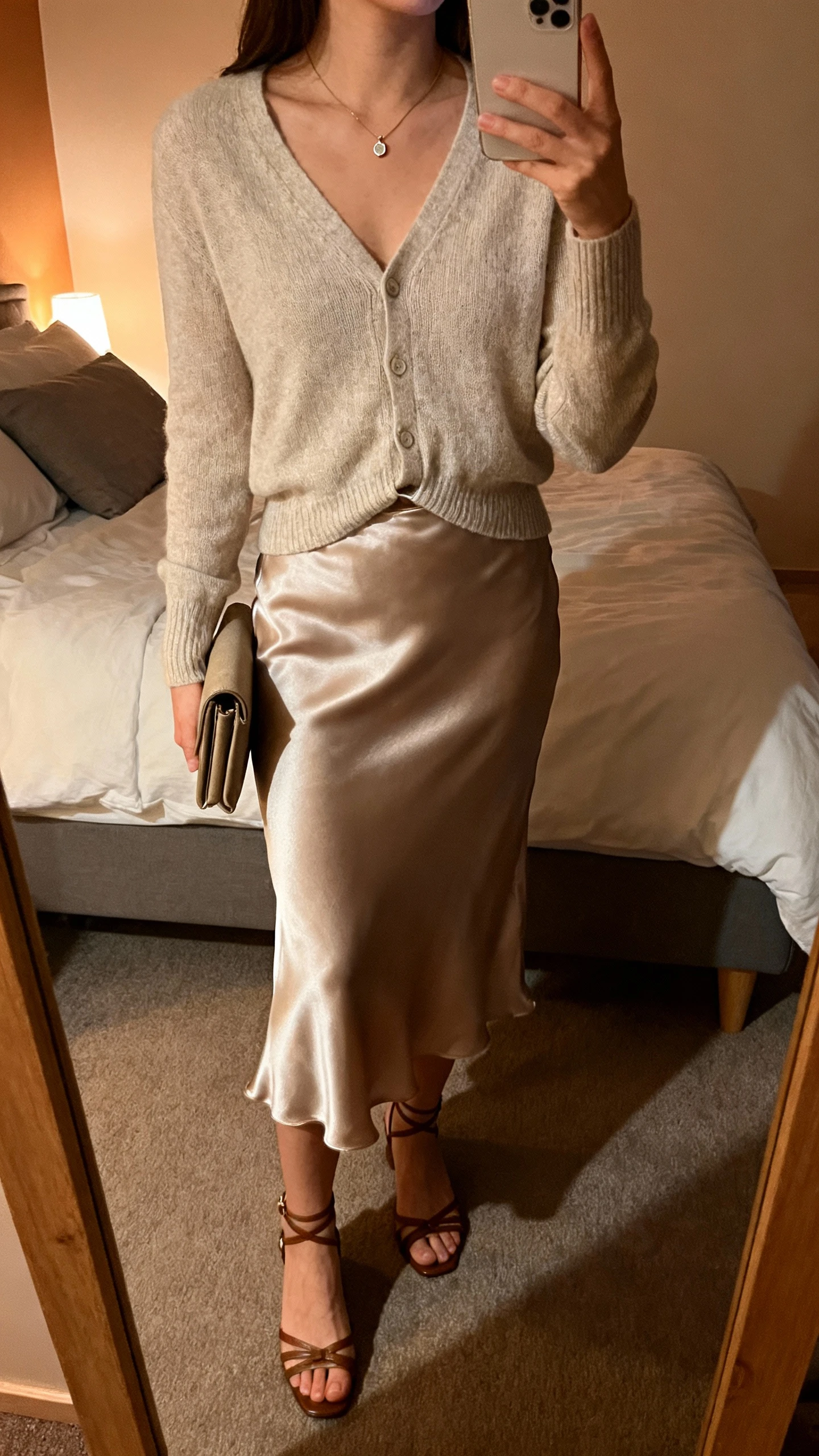 iPhone mirror selfie of a woman wearing a soft cardigan slightly tucked into a satin slip skirt, strappy low heels, a petite pendant necklace, and a clutch, face not visible, cropped at neck, bedroom mirror, warm indoor evening light, iPhone photo quality.