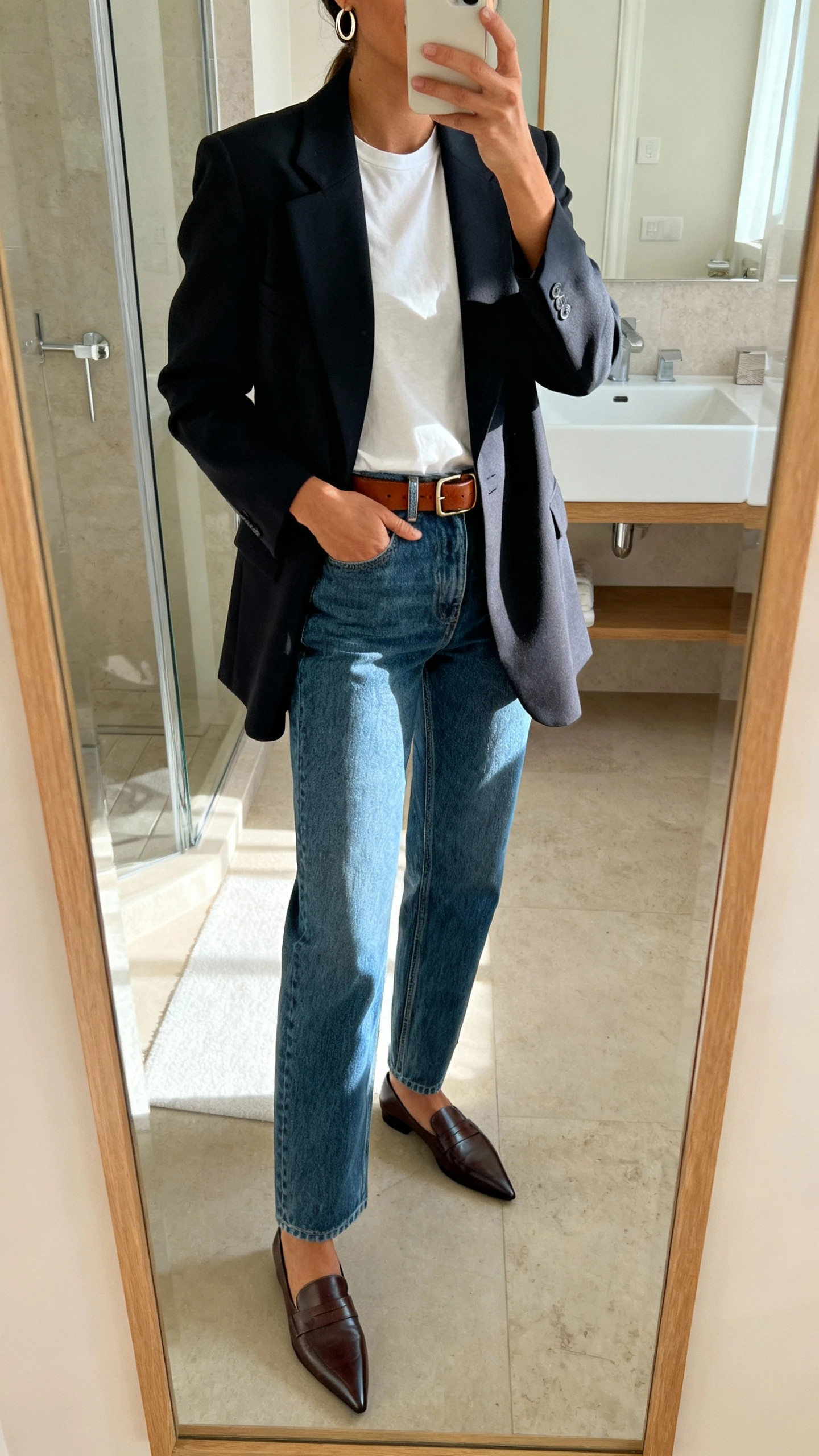 iPhone mirror selfie of a woman wearing a structured blazer over a simple tee, straight-leg denim, pointed loafers, and a leather belt with minimal hoop earrings, face not visible, bathroom full-length mirror, indoor daylight, iPhone photo quality.