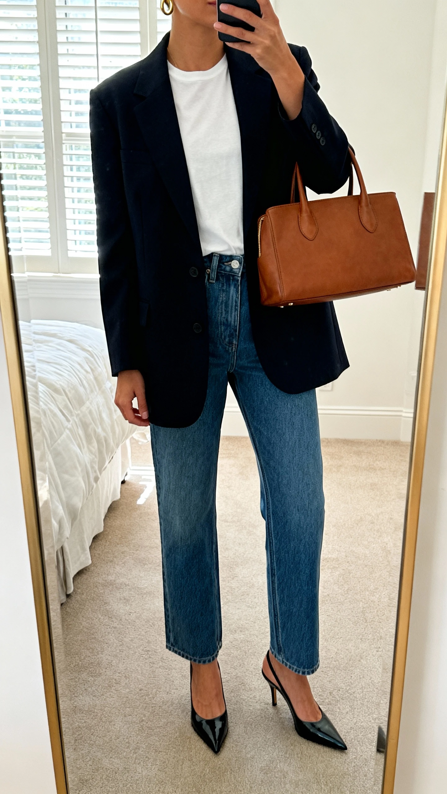 iPhone mirror selfie of a woman wearing a tailored blazer over a white tee with elevated straight-leg denim, pointed-toe heels, a structured leather tote, and minimal gold hoops, face not visible, cropped at neck, bedroom full-length mirror, natural window light, iPhone photo quality.