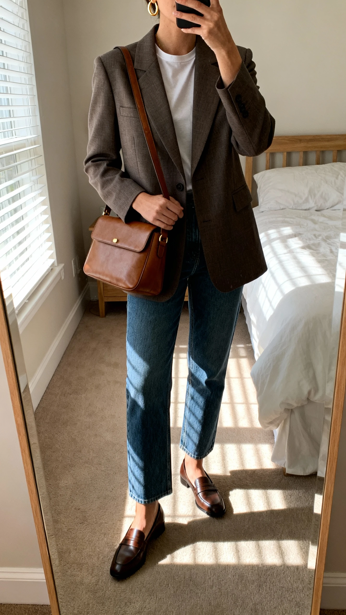 iPhone mirror selfie of a woman wearing a tailored blazer over a simple tee, straight-leg denim, low-heeled loafers, a structured leather shoulder bag, and minimalist gold hoops, face not visible, cropped at neck, bedroom mirror with morning window light, iPhone photo quality.