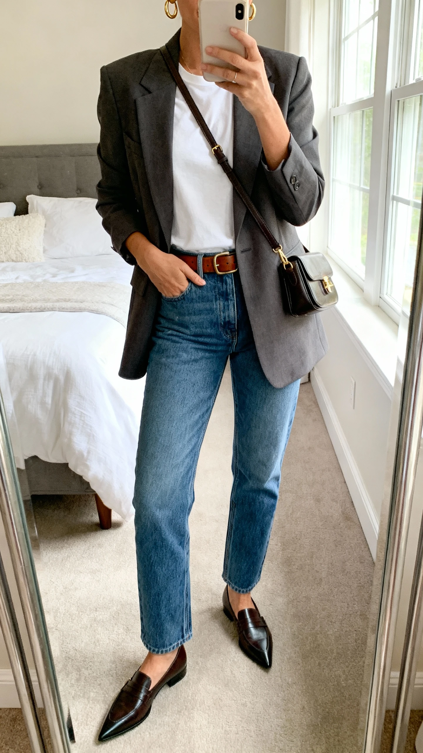 iPhone mirror selfie of a woman wearing a tailored blazer over a crisp white tee, straight-leg blue jeans, leather belt, pointed-toe loafers, structured crossbody bag, and minimalist gold hoops, face not visible, cropped at neck, bedroom mirror, morning window light, iPhone photo quality.