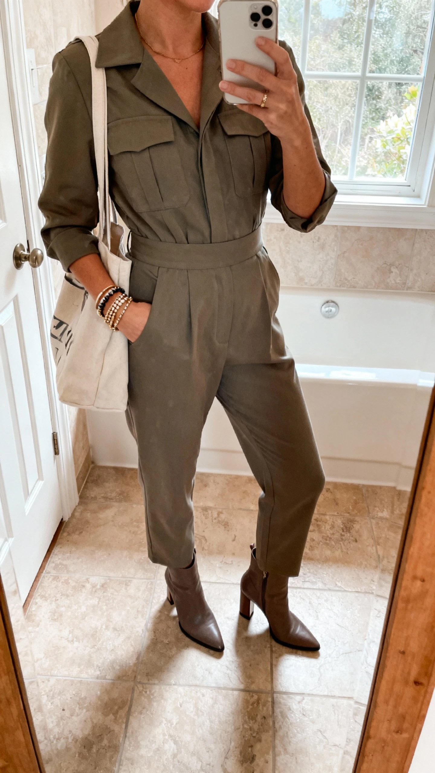 iPhone mirror selfie of a woman wearing a tailored utility-style jumpsuit with a defined waist, heeled ankle boots, layered bracelets, and a tote bag, face not visible, cropped at neck, bathroom mirror, natural window light, casual iPhone photo.