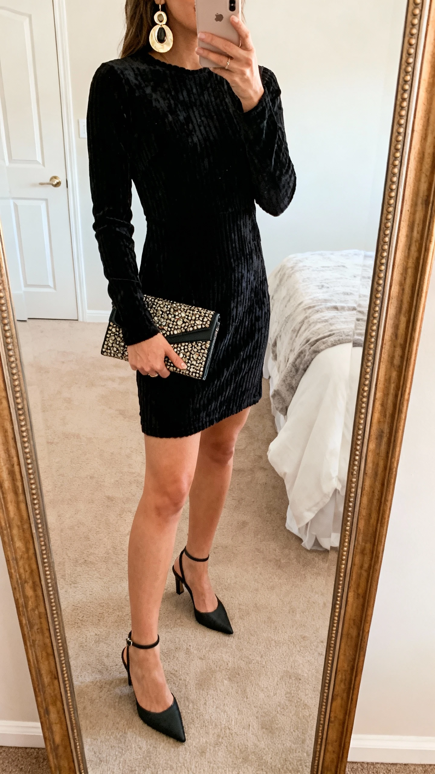 iPhone mirror selfie of a woman wearing a textured little black dress (think ribbed or velvet) with slingback heels, a statement clutch, and bold earrings, face not visible, cropped at shoulders, bedroom mirror, warm indoor lighting, iPhone photo quality.