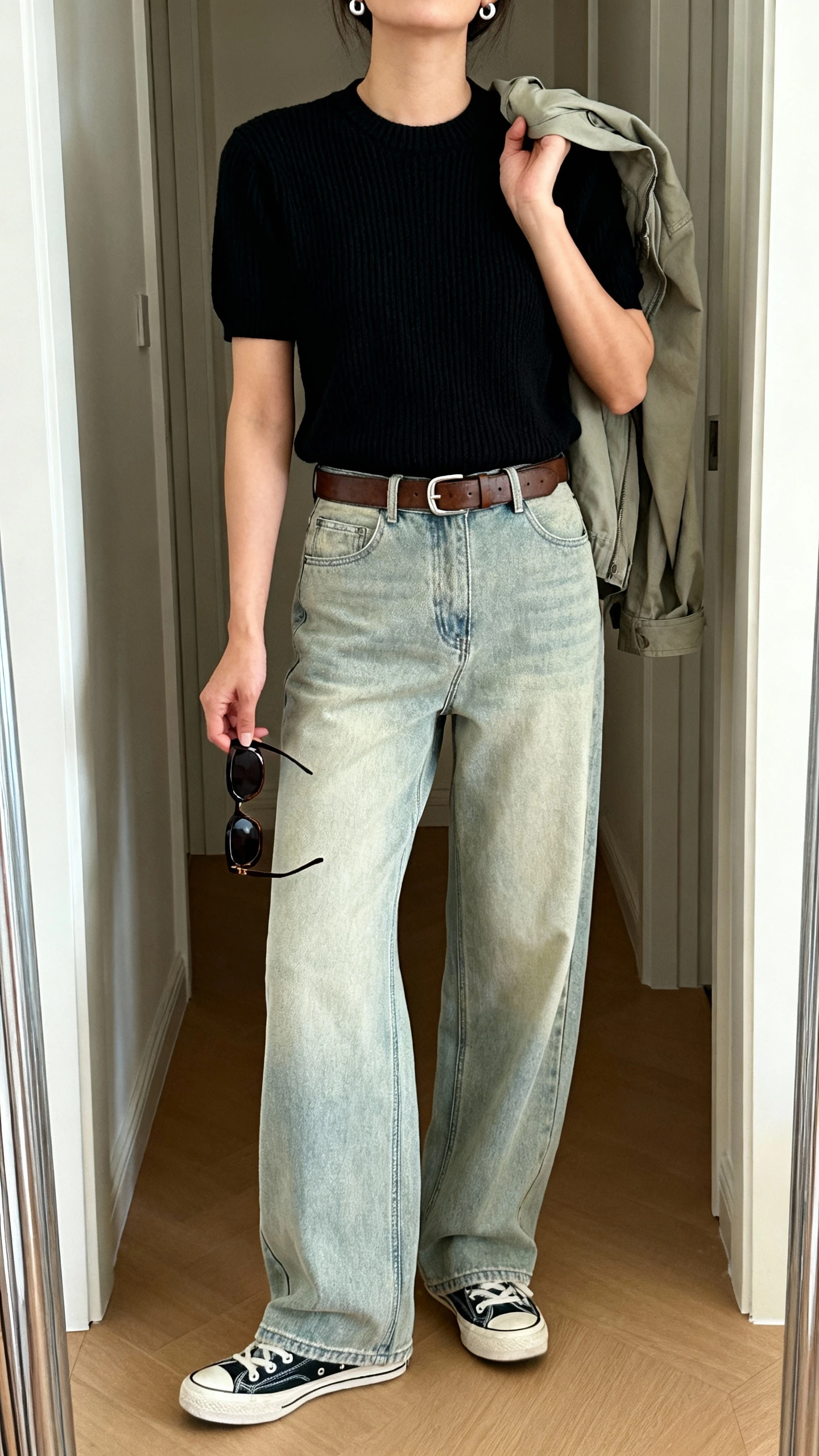iPhone mirror selfie of a woman wearing a thick-knit black crewneck tee, high-rise light-wash wide-leg jeans, clean retro sneakers, simple leather belt with minimal hardware, chunky sunglasses in hand and stud earrings, lightweight utility jacket thrown over shoulder; face not visible, cropped at shoulders, full-length mirror in hallway with natural daylight, iPhone photo quality.