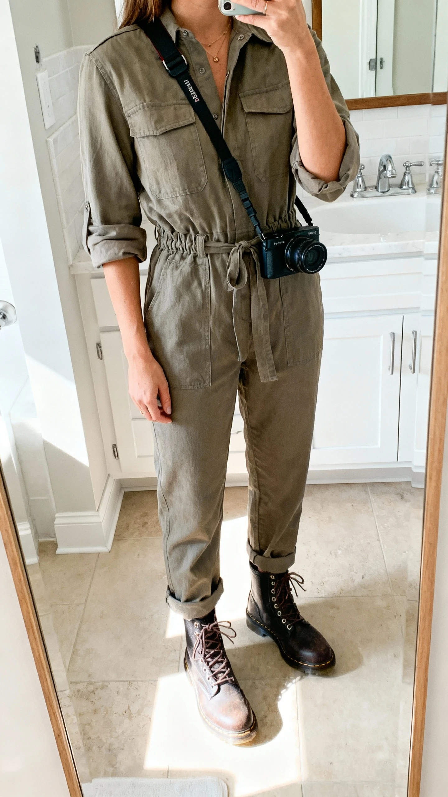 iPhone mirror selfie of a woman wearing a utility jumpsuit with rolled sleeves, cinched waist, lace-up boots, and a crossbody camera bag, face not visible, bathroom mirror, bright indoor light, iPhone photo quality.