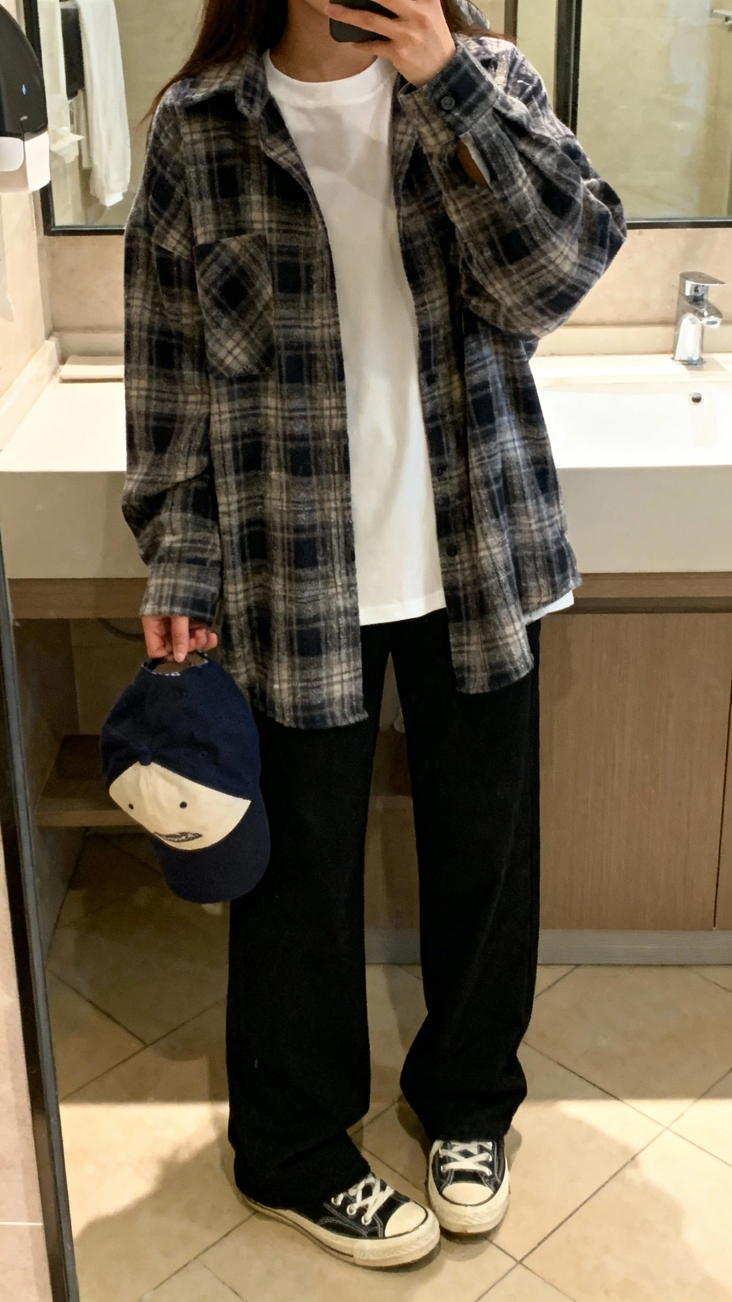 iPhone mirror selfie of a woman wearing a white crewneck tee layered under an oversized plaid flannel with straight-leg black pants, canvas sneakers, baseball cap in hand, face not visible, bathroom mirror with warm indoor lighting, iPhone photo quality
