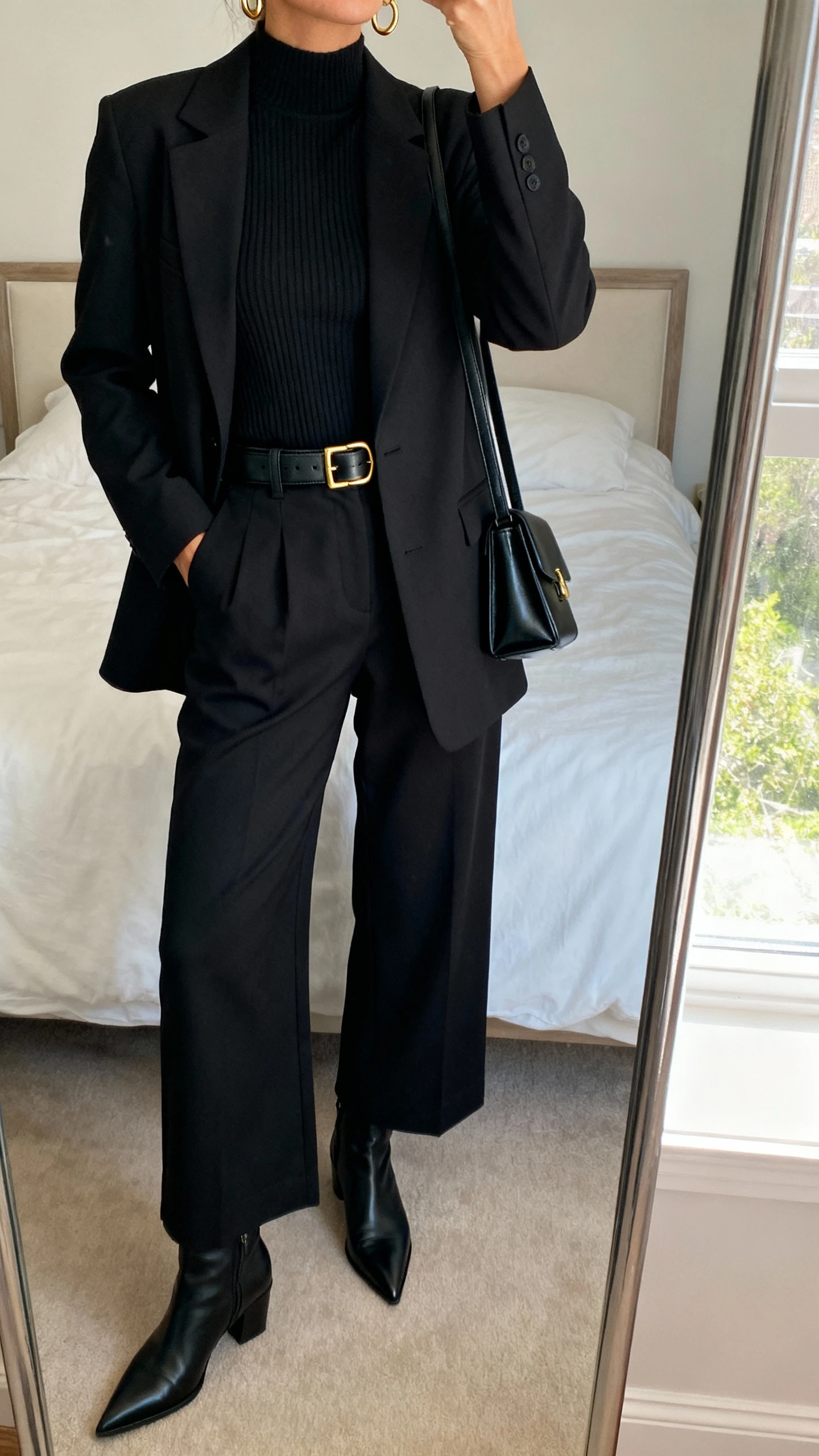 iPhone mirror selfie of a woman wearing an all-black elevated monochrome look: black tailored blazer, black ribbed mock-neck top, black high-waisted wide-leg trousers, black leather belt with subtle gold buckle, black pointed-toe ankle boots, small black structured shoulder bag, delicate gold hoop earrings, face not visible, cropped at neck, bedroom mirror with morning window light, iPhone photo quality