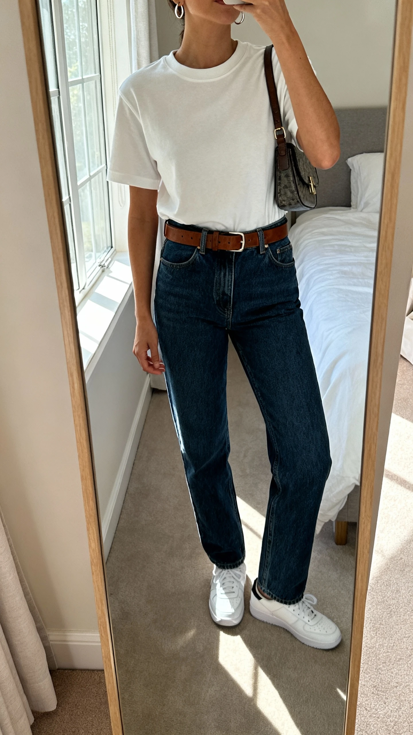 iPhone mirror selfie of a woman wearing an elevated crewneck tee tucked into high-rise straight-leg jeans with a leather belt, sleek white sneakers, small hoop earrings, and a structured crossbody bag, face not visible, cropped at neck, bedroom mirror, natural window light, iPhone photo quality