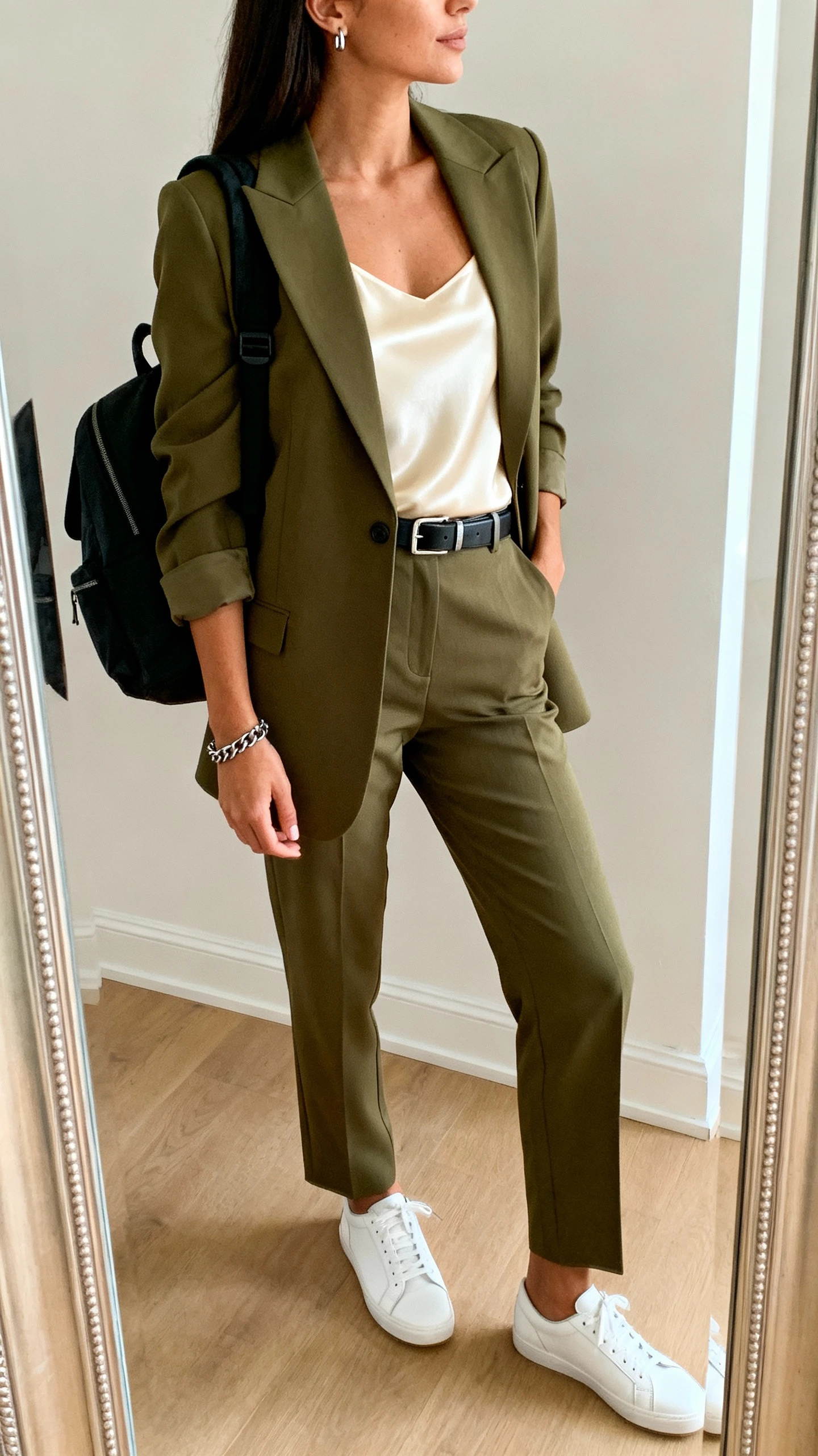 iPhone mirror selfie of a woman wearing an olive well-cut blazer with matching straight-leg trousers, a silky cream camisole, clean white leather sneakers, a sleek belt, modern stud earrings, a chain bracelet, and an understated black backpack, face not visible, side angle in full-length mirror, indoor daylight, iPhone photo quality.