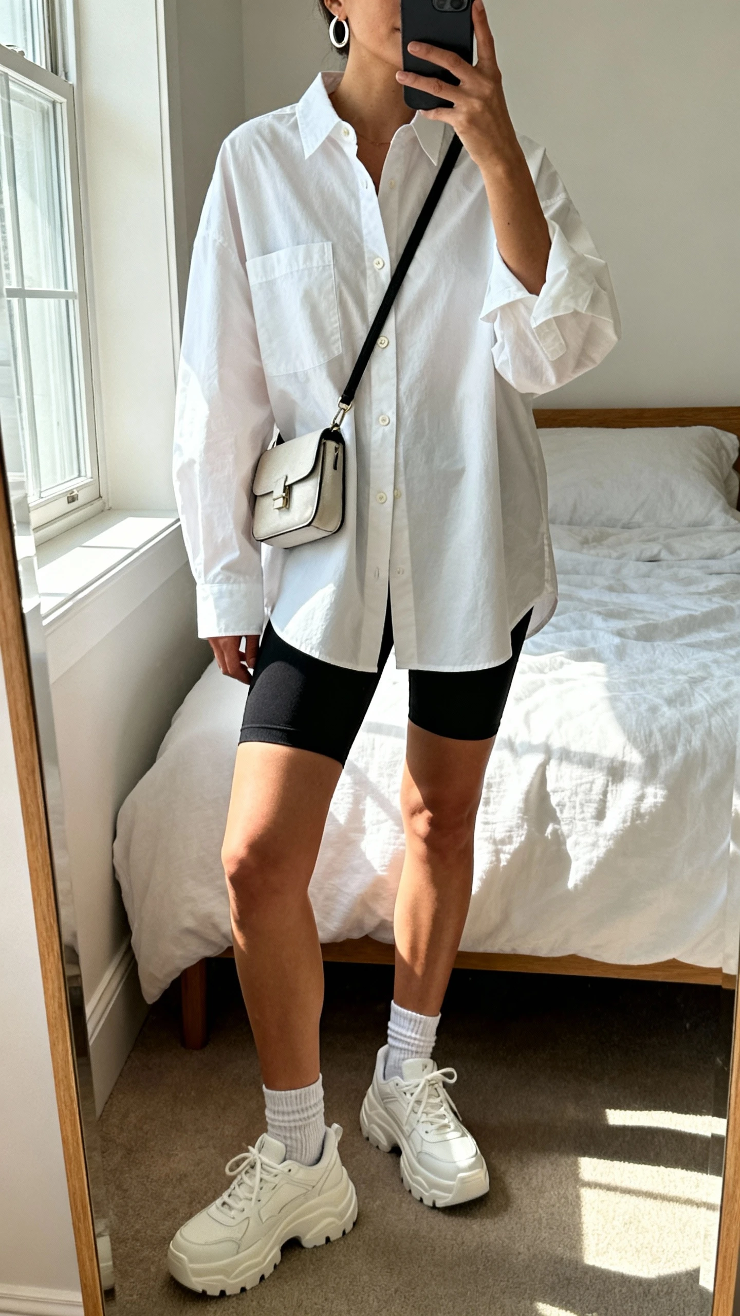 iPhone mirror selfie of a woman wearing an oversized white button-up shirt with black bike shorts, ankle socks, chunky white sneakers, small crossbody bag, minimalist hoop earrings, face not visible, cropped at neck, bedroom mirror with natural morning window light, iPhone photo quality