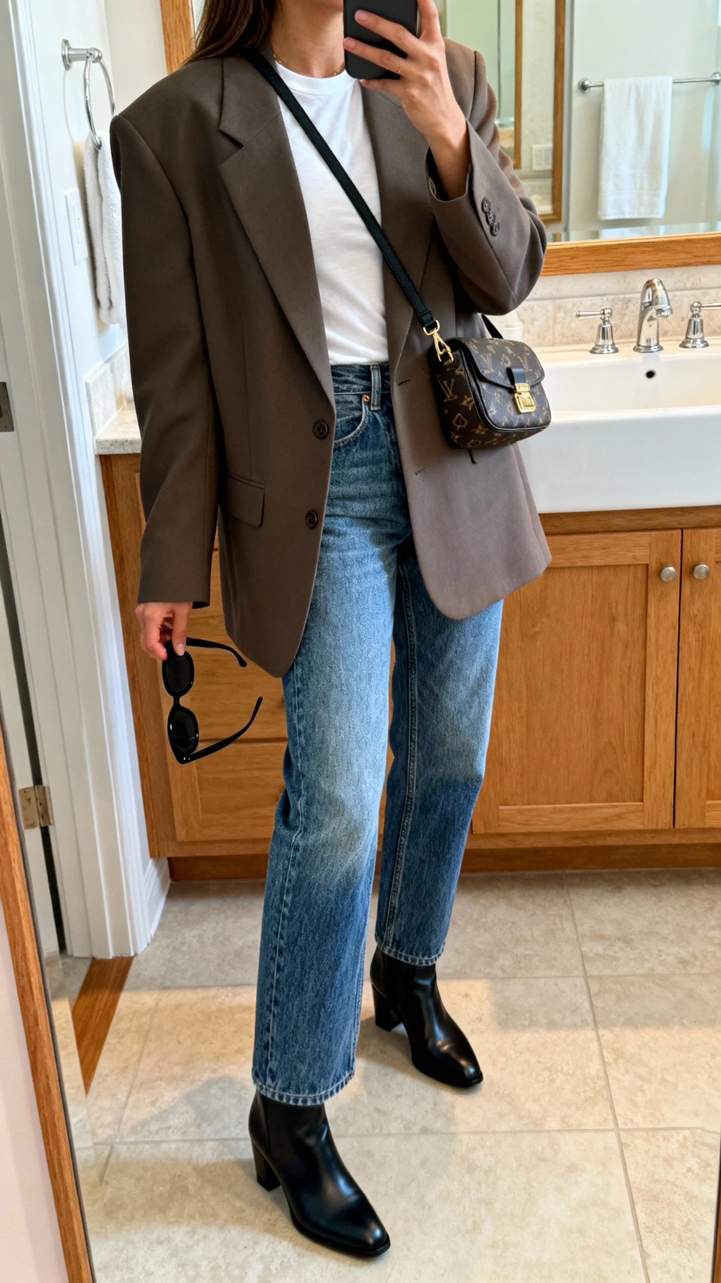 iPhone mirror selfie of a woman wearing an oversized blazer over a fitted white tee with straight-leg vintage blue jeans, sleek ankle boots, designer-looking crossbody bag, and black sunglasses in hand, face not visible, side angle, bathroom mirror, morning indoor light, iPhone photo quality.