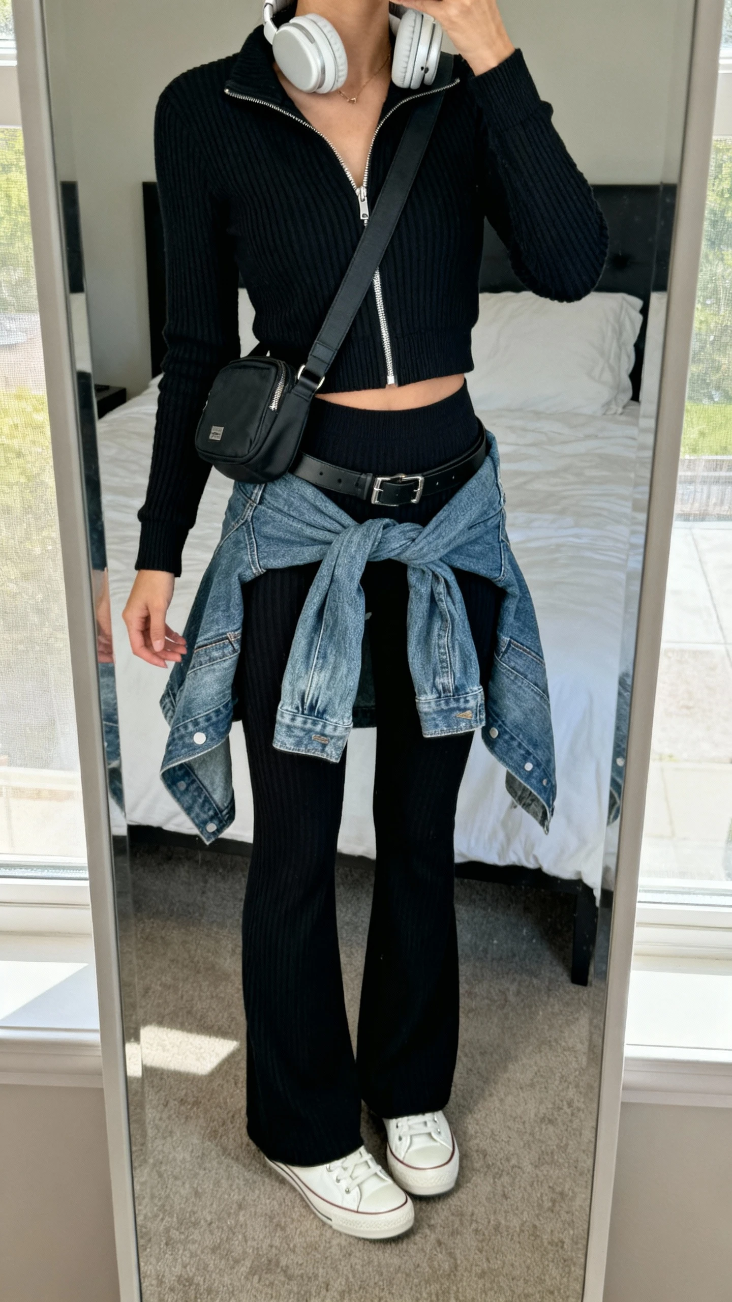iPhone mirror selfie of a woman wearing elevated airport athleisure: matching black ribbed knit set (zip-up track jacket and high-waist flare leggings), white leather sneakers, oversized denim jacket tied at waist, crossbody belt bag, over-ear headphones, face not visible, cropped at neck, bedroom mirror, natural window light, iPhone photo quality.
