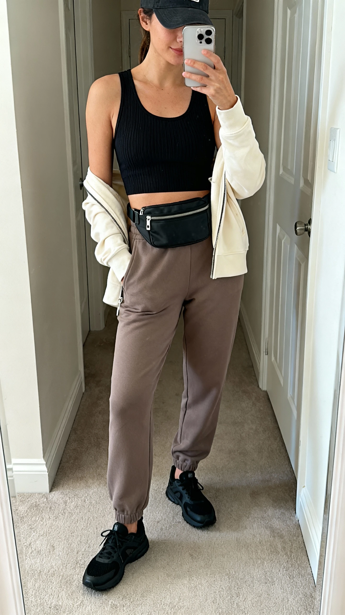 iPhone mirror selfie of a woman wearing elevated athleisure: black ribbed sports tank, tailored taupe joggers, lightweight cream zip-up, sleek black trainers, baseball cap, crossbody belt bag worn across chest, face not visible, cropped at neck, entryway full-length mirror with natural light, iPhone photo quality.