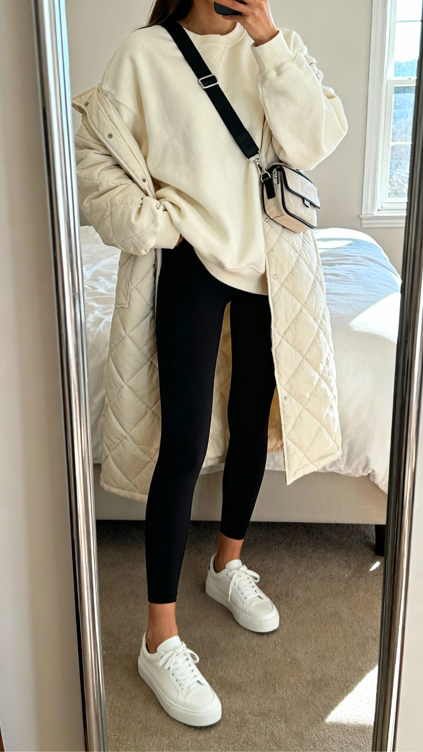 iPhone mirror selfie of a woman wearing elevated black leggings, an oversized cream sweatshirt, a longline quilted jacket, white low-profile sneakers, and a crossbody belt bag, face not visible, cropped at neck, bedroom mirror with morning window light, iPhone photo quality.