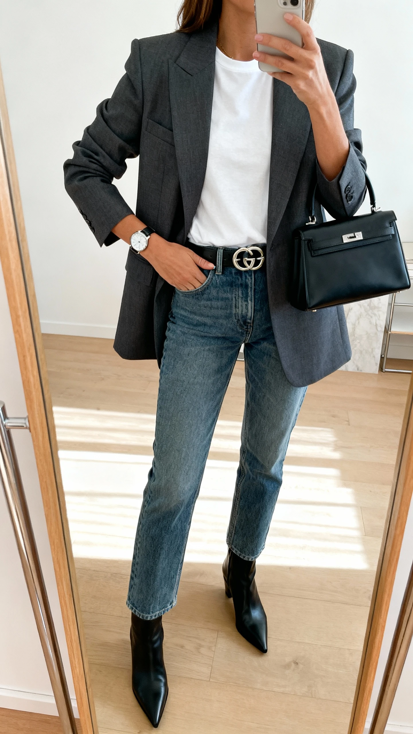 iPhone mirror selfie of a woman wearing elevated denim with a power blazer: structured charcoal blazer over a crisp white tee, medium-wash straight jeans, pointed black ankle boots, statement belt and sleek watch, small top-handle bag, face not visible, full-length mirror, bright indoor lighting, iPhone photo quality.