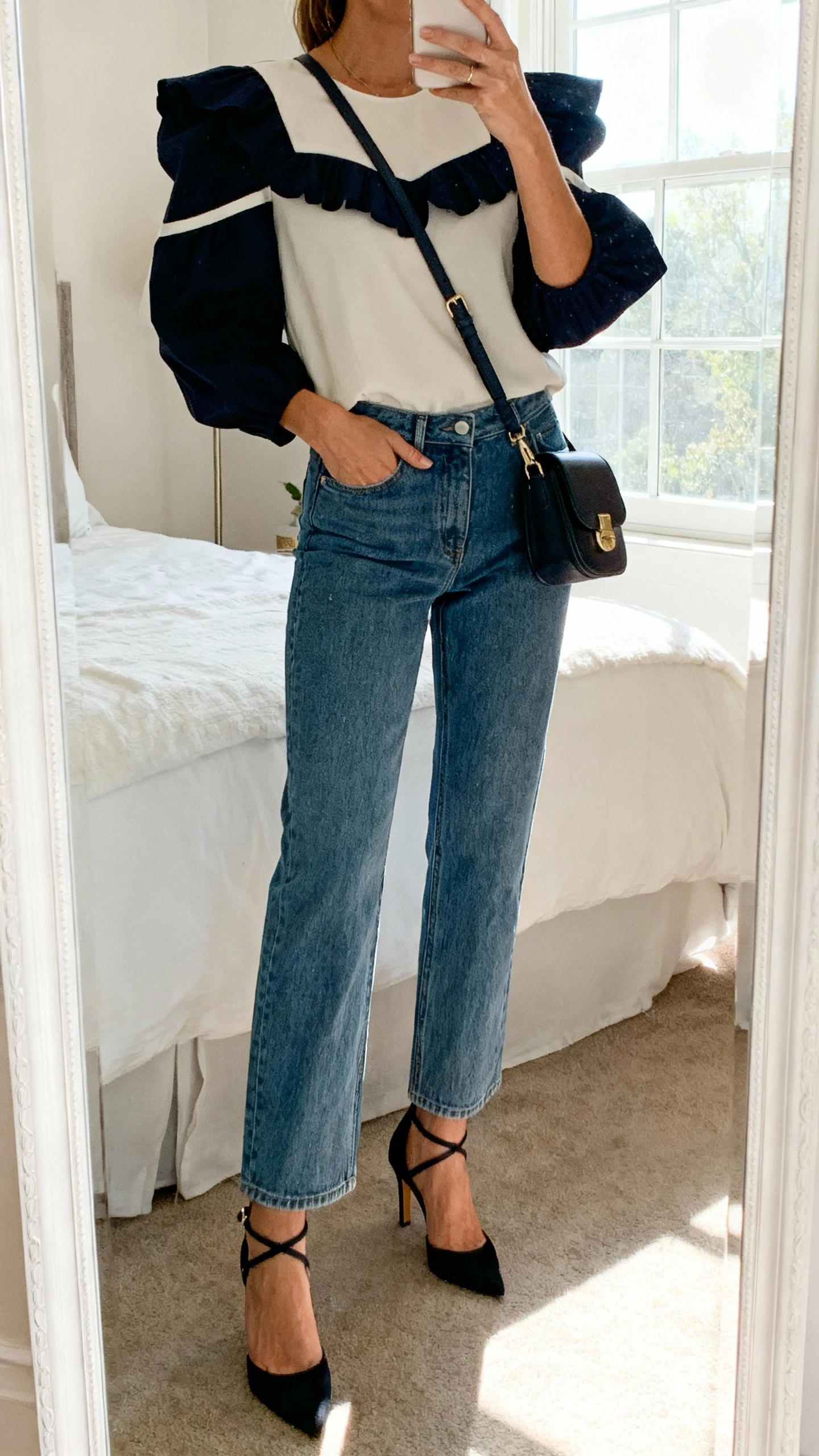 iPhone mirror selfie of a woman wearing elevated straight-leg jeans, a statement top (bold sleeves or interesting neckline), ankle-strap heels, and a crossbody bag, face not visible, cropped at shoulders, bedroom mirror, natural window light, iPhone photo quality.