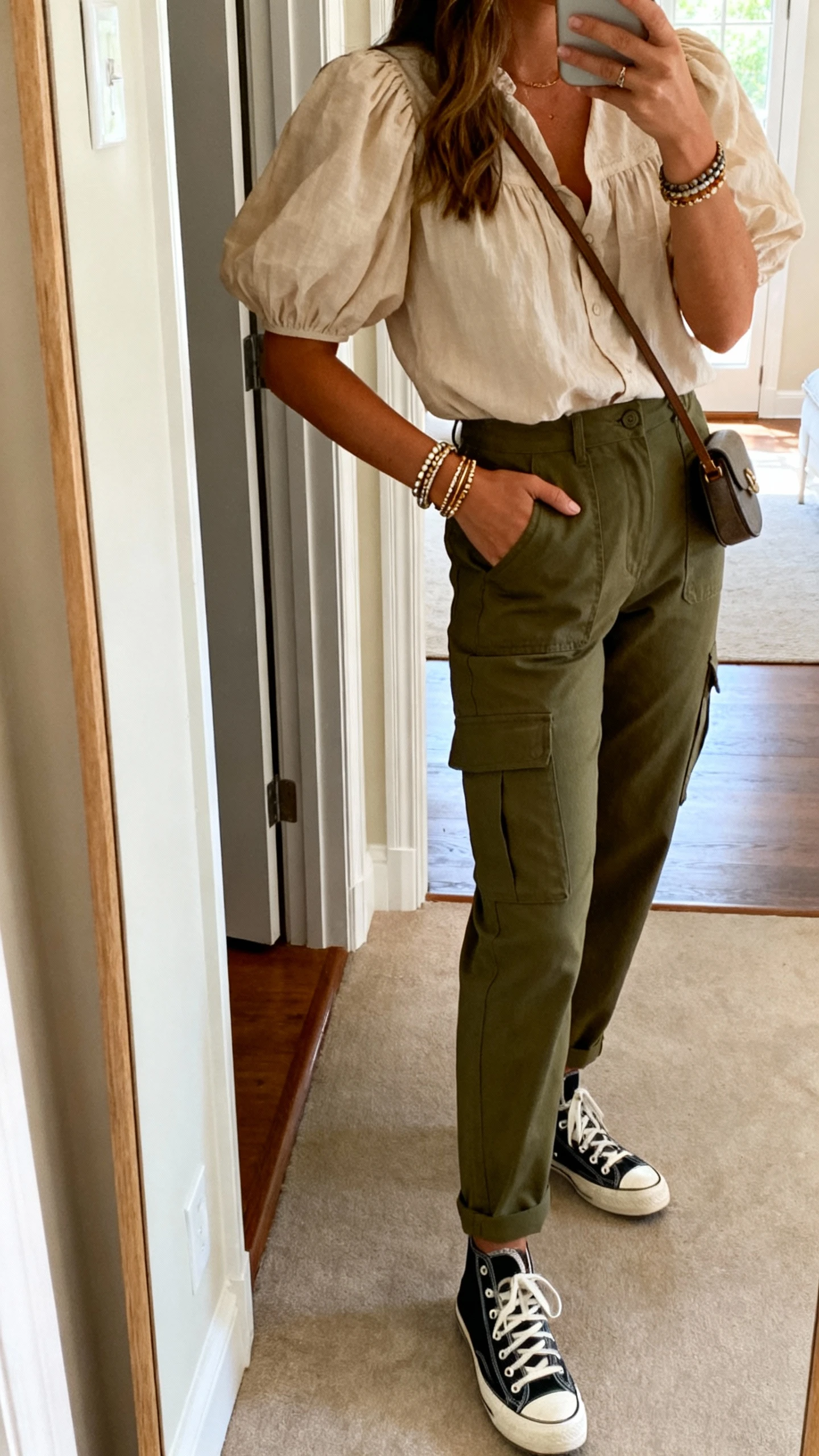 iPhone mirror selfie of a woman wearing olive utility cargo pants with a feminine puff-sleeve blouse, stacked bracelets, low-top canvas sneakers, and a small crossbody, face not visible, side angle, entryway mirror, natural morning light, iPhone photo quality
