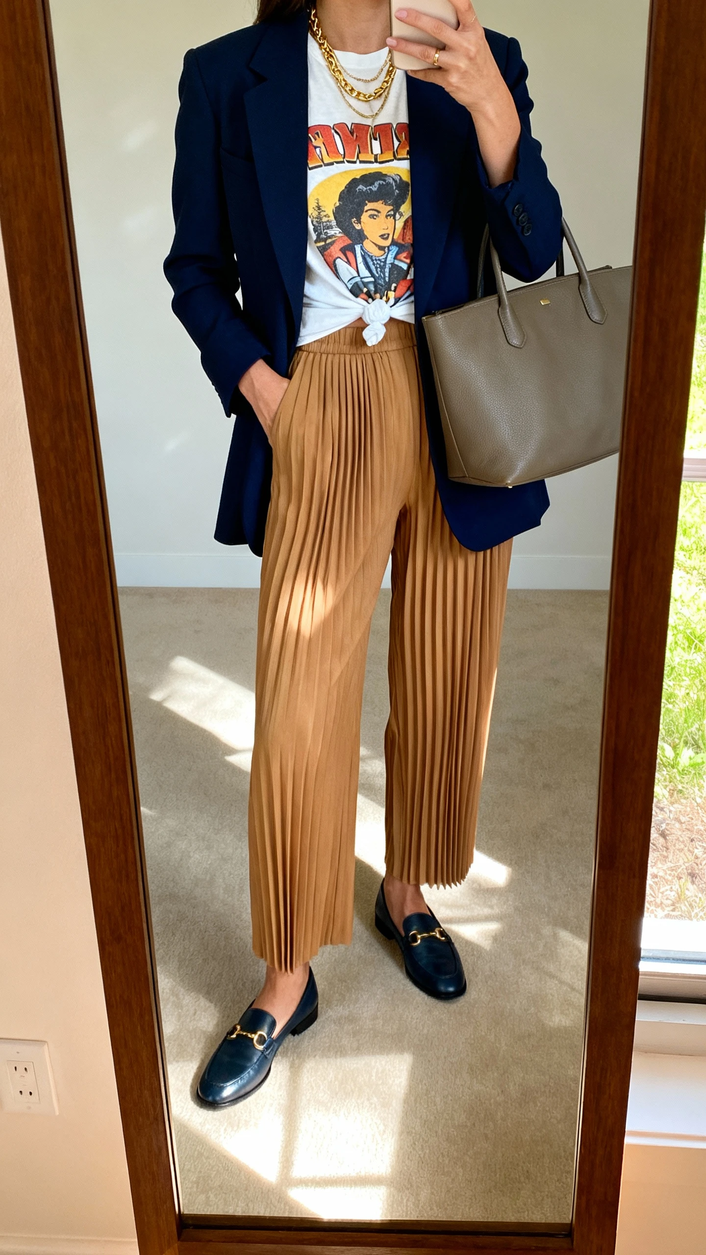 iPhone mirror selfie of a woman wearing pleated high-waisted trousers in camel, vintage graphic tee half-tucked, navy blazer, loafers, layered gold necklaces, and a structured tote, face not visible, cropped at shoulders, full-length mirror, soft afternoon indoor lighting, iPhone photo quality.