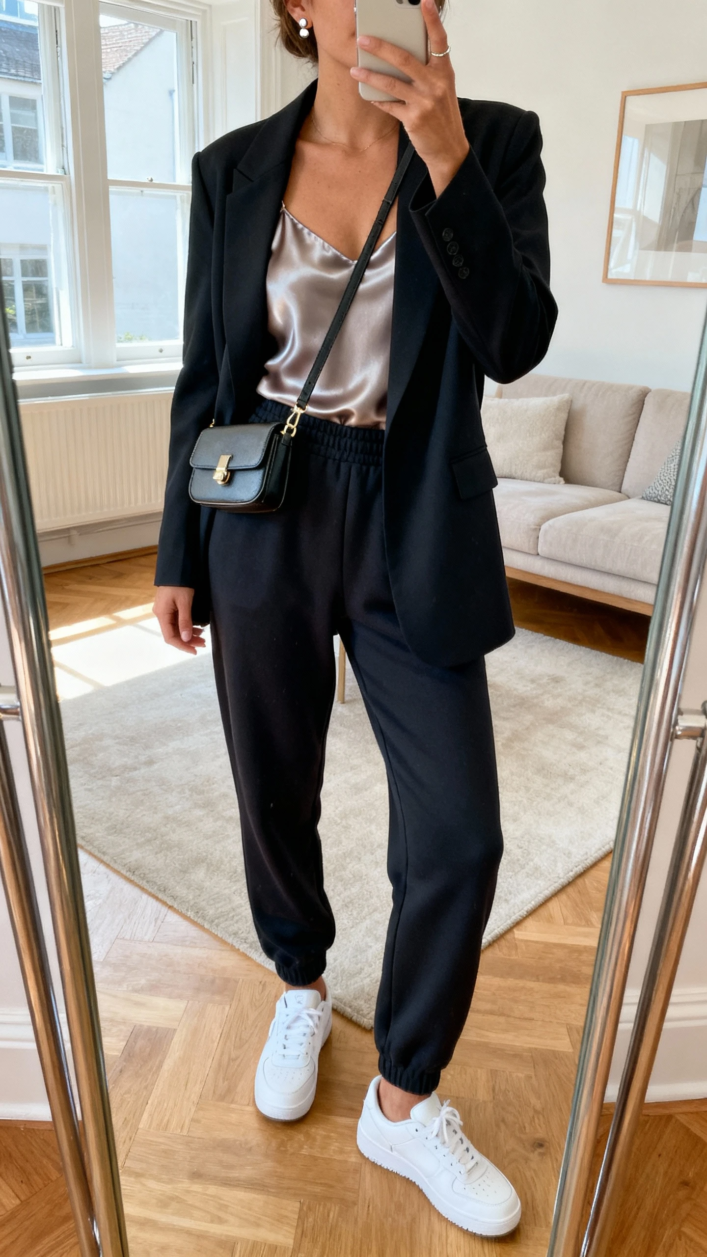 iPhone mirror selfie of a woman wearing tailored joggers with a silky camisole and a sleek blazer, clean white sneakers, small crossbody, and simple studs, face not visible, cropped at shoulders, living room full-length mirror, natural window light, iPhone photo quality.