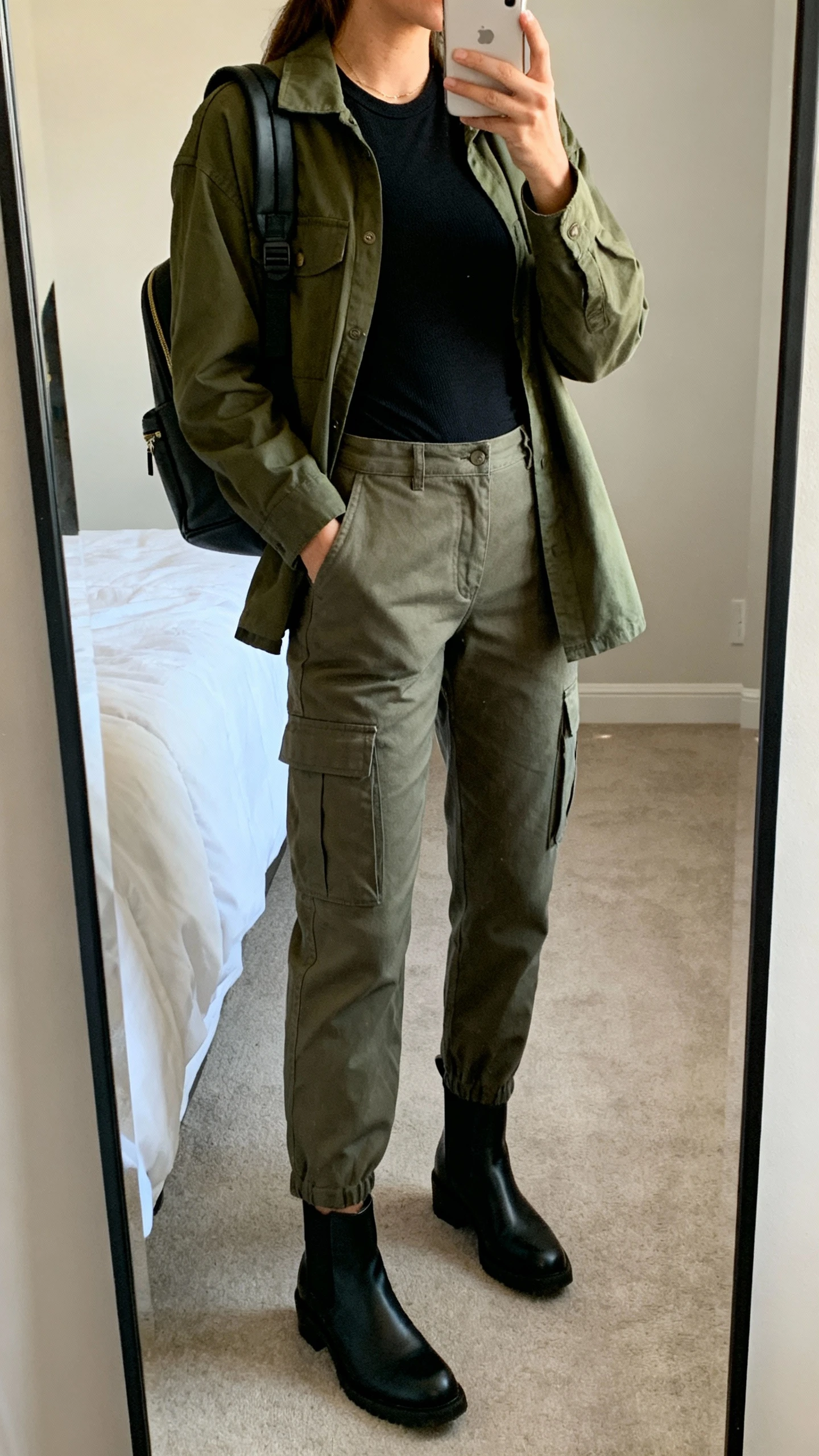 iPhone mirror selfie of a woman wearing utility-inspired cargo pants, a fitted black tee, an olive chore jacket, black ankle Chelsea boots, and a structured backpack, face not visible, cropped at neck, bedroom mirror with soft indoor lighting, iPhone photo quality.