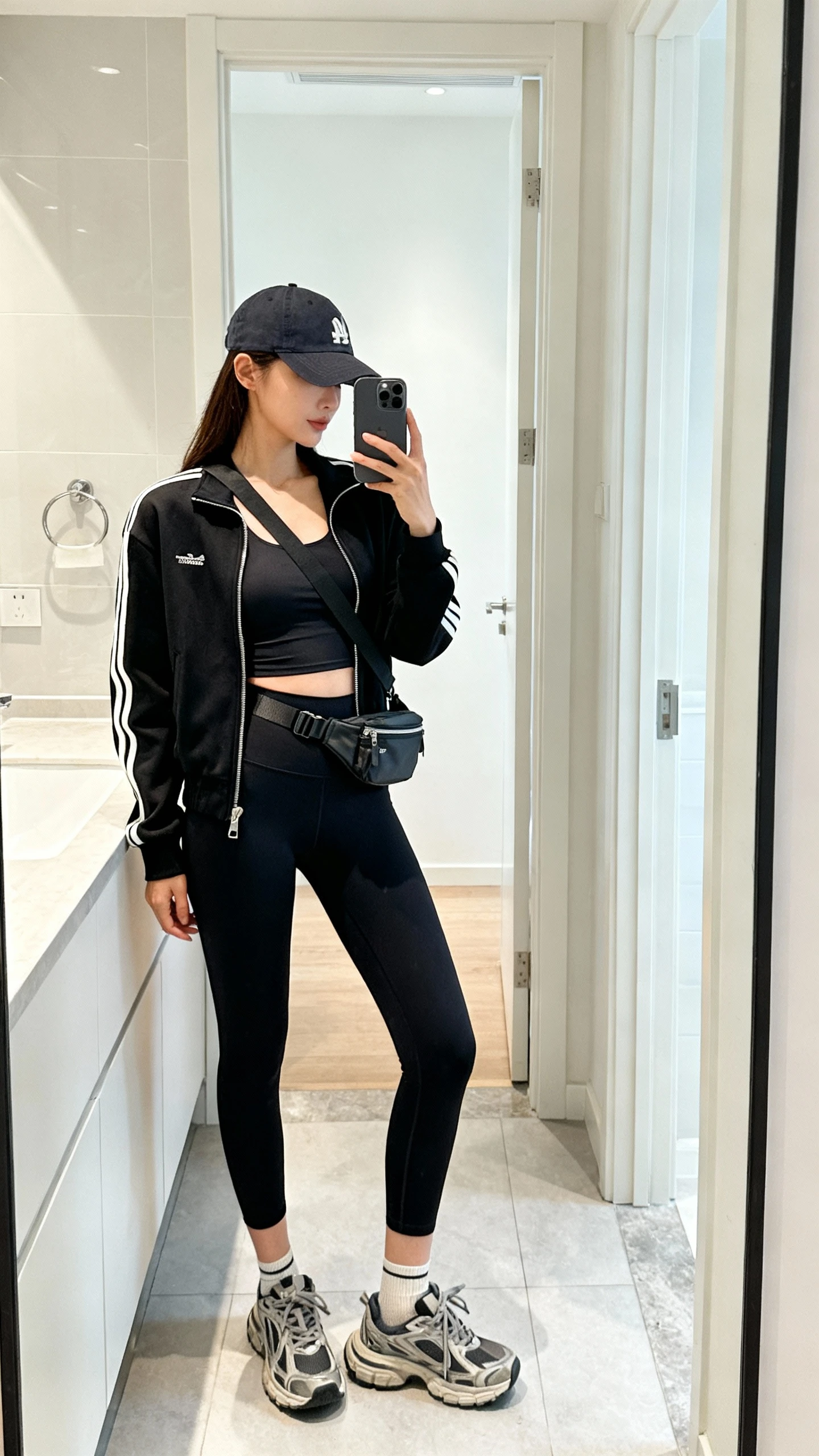 iPhone side-angle mirror selfie of a woman in a black zip-up track jacket over a fitted tank, high-waisted black leggings, chunky retro sneakers, a baseball cap, crossbody belt bag, and thin athletic socks, face not visible, bathroom full-length mirror, bright indoor lighting, iPhone photo quality.