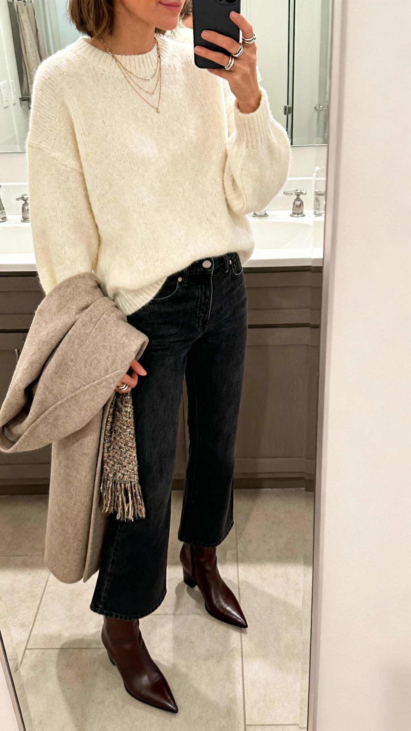 iPhone side-angle mirror selfie of a woman in a boxy chunky cream sweater, high-rise dark-wash cropped flare jeans, pointed-toe leather ankle boots, layered delicate necklaces and stacked rings, with a cozy wool coat draped over arm and a textured scarf; face not visible, bathroom mirror, soft indoor lighting, iPhone photo quality.