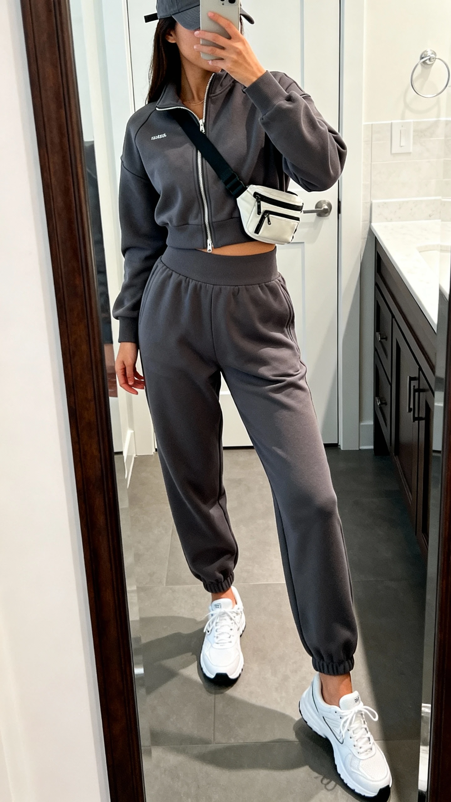 iPhone side-angle mirror selfie of a woman in a matching sporty set: charcoal zip-up track jacket and high-waist joggers with white athletic sneakers, baseball cap, small crossbody belt bag, face not visible, bathroom full-length mirror, bright indoor lighting, iPhone photo quality.