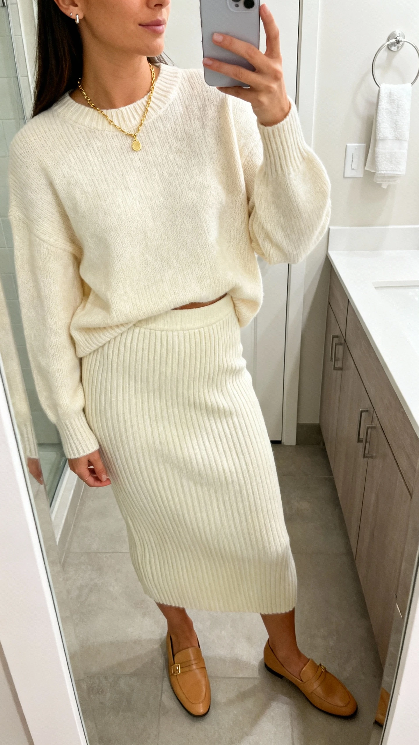 iPhone side-angle mirror selfie of a woman in a monochrome knit set: matching cream ribbed sweater and high-waisted ribbed knit skirt, delicate gold necklace, simple stud earrings, and sleek tan loafers; face not visible, bathroom mirror with soft indoor lighting, iPhone photo quality.