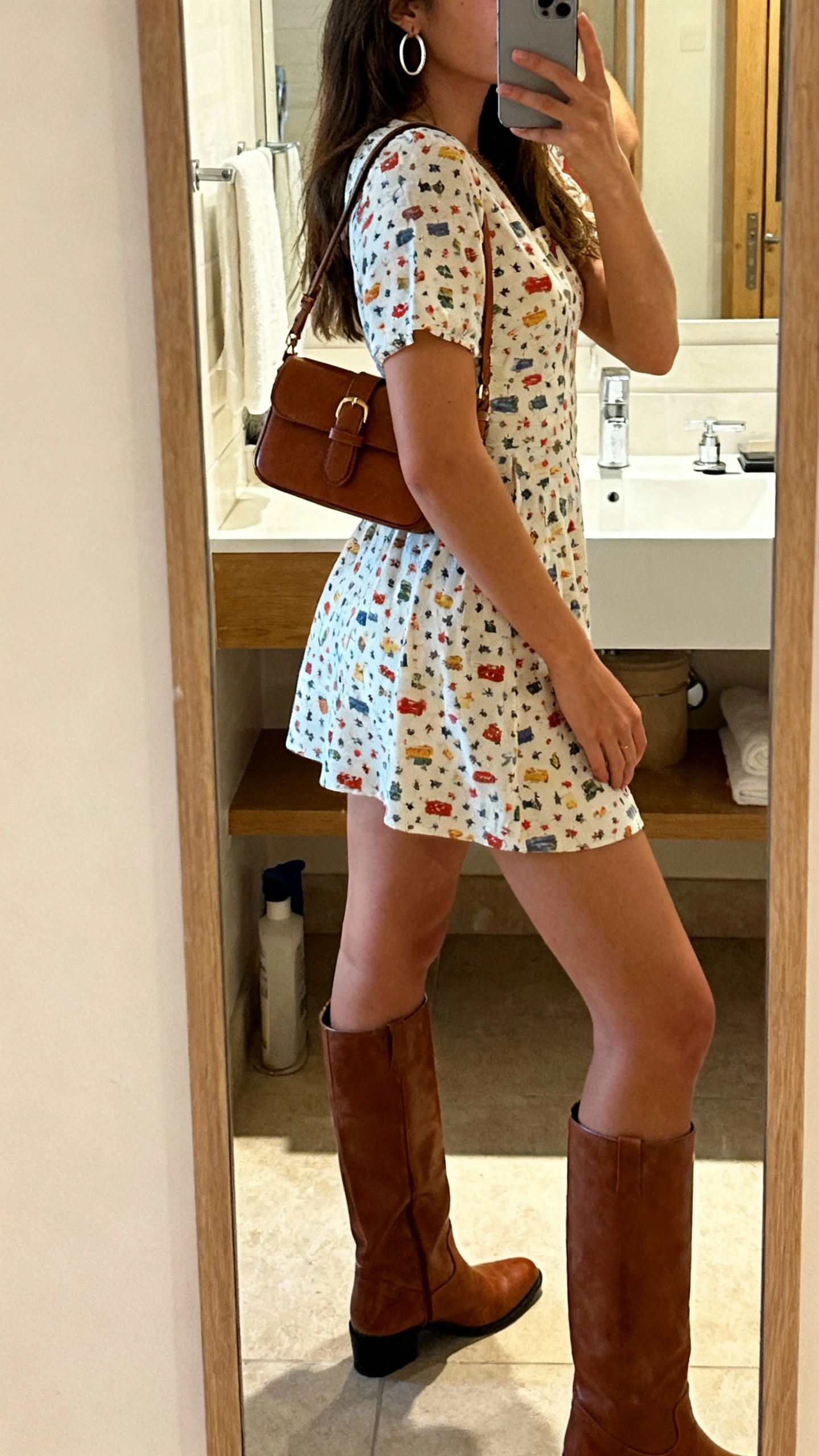 iPhone side-angle mirror selfie of a woman in a playful mini dress with knee-high boots, small shoulder bag, and hoop earrings, face not visible, bathroom full-length mirror, indoor warm lighting, iPhone photo quality.