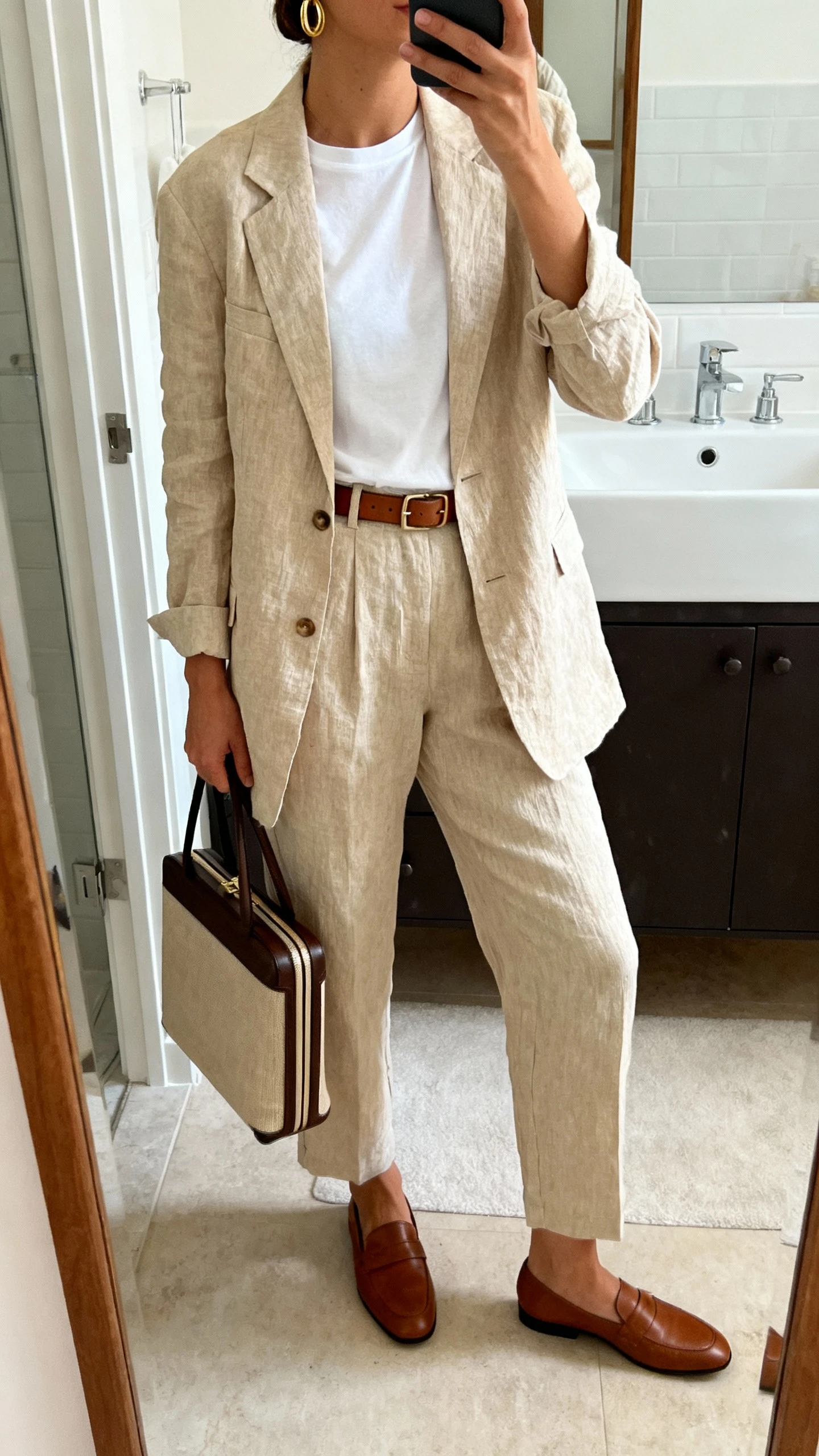 iPhone side-angle mirror selfie of a woman wearing a beige linen blazer over a white crewneck tee, tailored linen trousers, slim leather belt, minimalist gold hoops, structured carry-on tote, brown loafers, face not visible, bathroom full-length mirror, morning indoor light, iPhone photo quality.