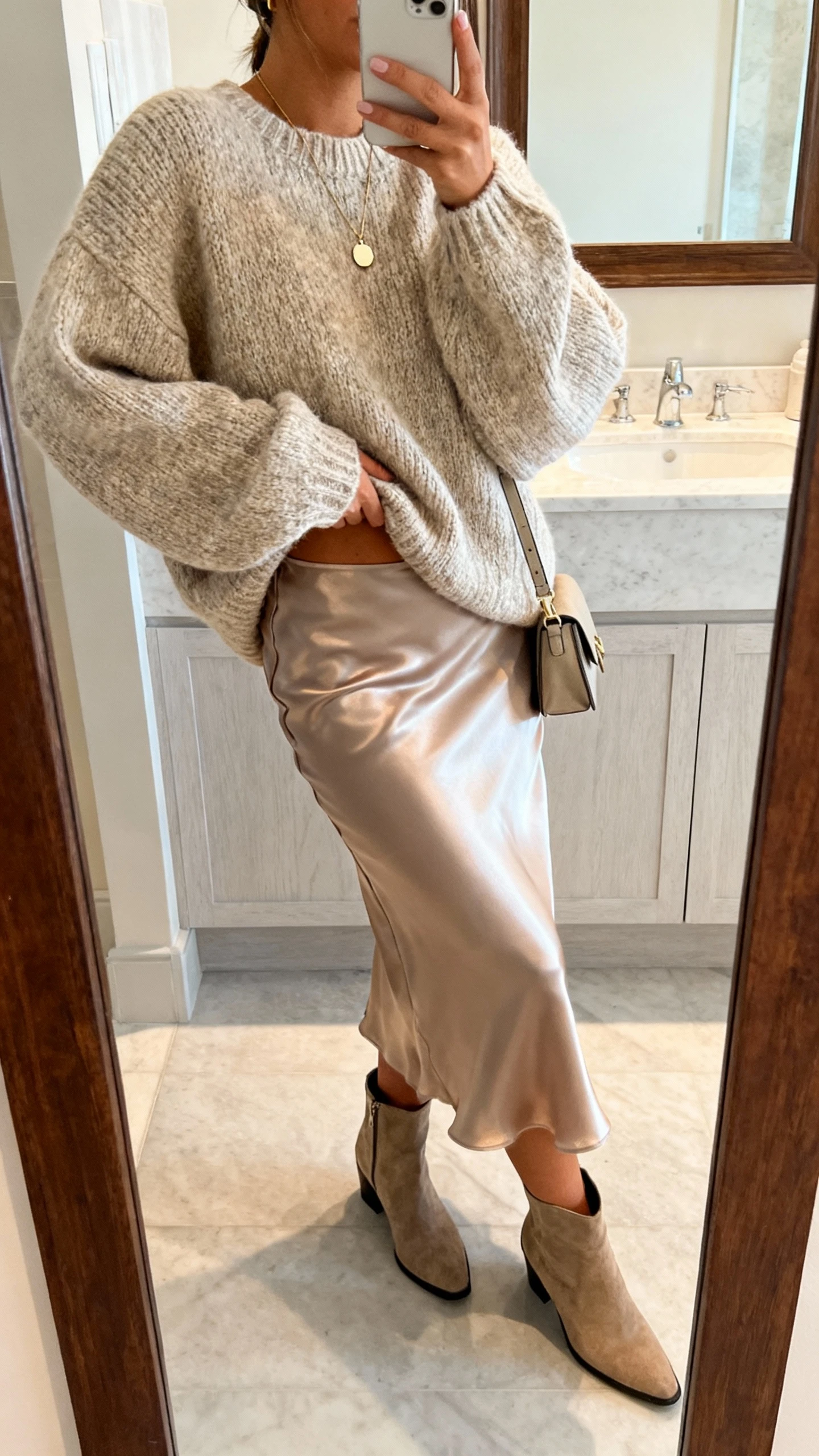 iPhone side-angle mirror selfie of a woman wearing a satin slip skirt, chunky oversized knit sweater half-tucked, ankle boots, delicate pendant necklace, and a small crossbody bag, face not visible, bathroom mirror with soft indoor lighting, iPhone photo quality.