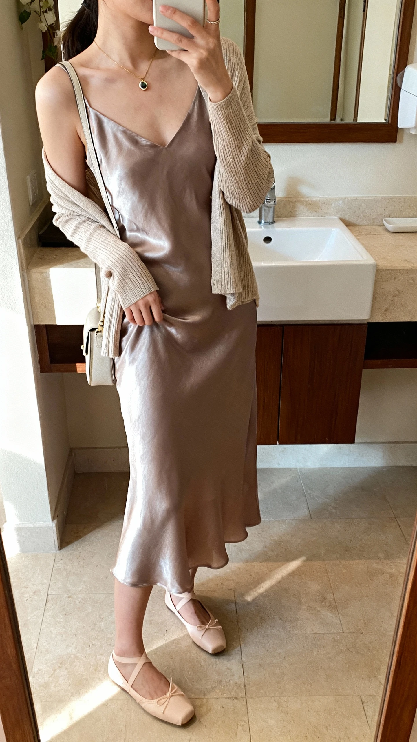 iPhone side-angle mirror selfie of a woman wearing a satin slip dress layered with a fine cardigan draped over shoulders, classic ballet flats, delicate pendant necklace, and a small shoulder bag, face not visible, bathroom mirror, soft morning light, iPhone photo quality.