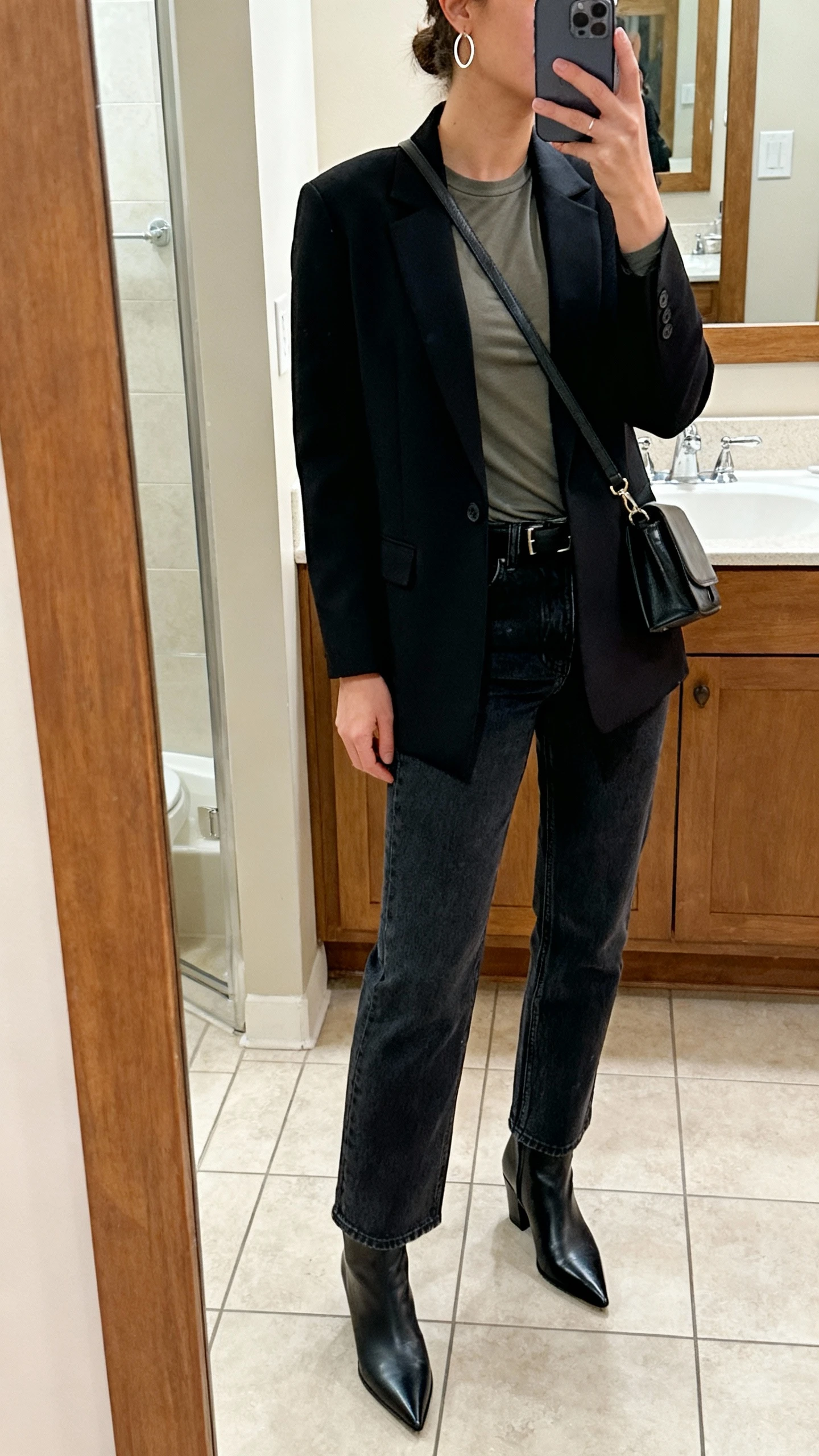 iPhone side-angle mirror selfie of a woman wearing straight-leg dark-wash jeans, a tailored blazer over a fitted tee, pointed-toe ankle boots, simple hoop earrings, and a structured crossbody, face not visible, bathroom mirror, soft indoor lighting, iPhone photo quality.