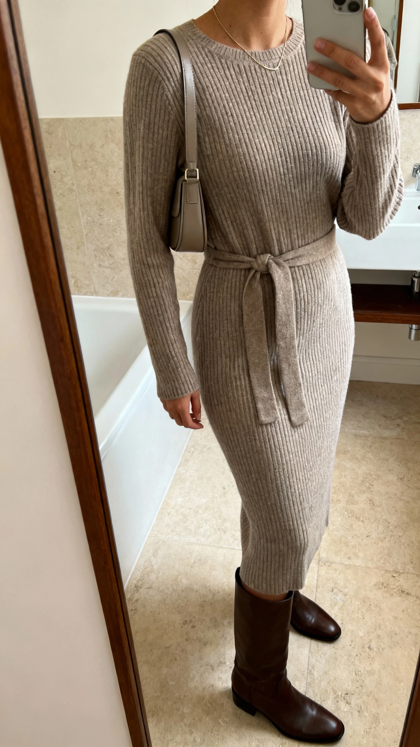 iPhone side-angle mirror selfie of a woman wearing a ribbed knit midi dress with a waist tie, knee-high leather boots, a slim shoulder bag, and delicate necklace, face not visible, cropped at shoulders, bathroom mirror, soft indoor lighting, iPhone photo quality.