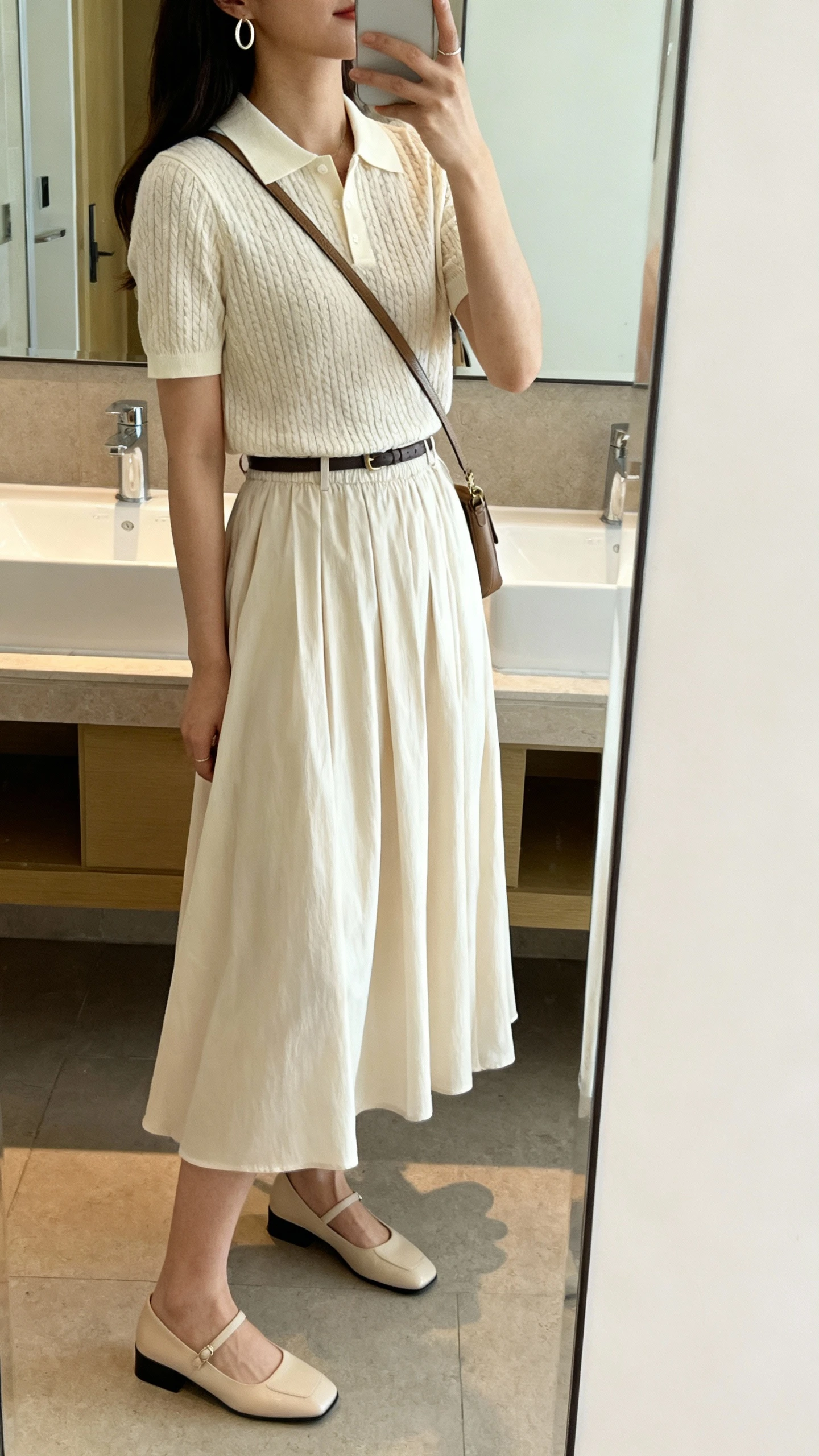 iPhone side-angle mirror selfie of a woman wearing a short-sleeve knit polo tucked into a flowy midi skirt, low block heels, minimal hoop earrings, and a crossbody bag, face not visible, bathroom mirror, soft indoor lighting, casual iPhone photo.