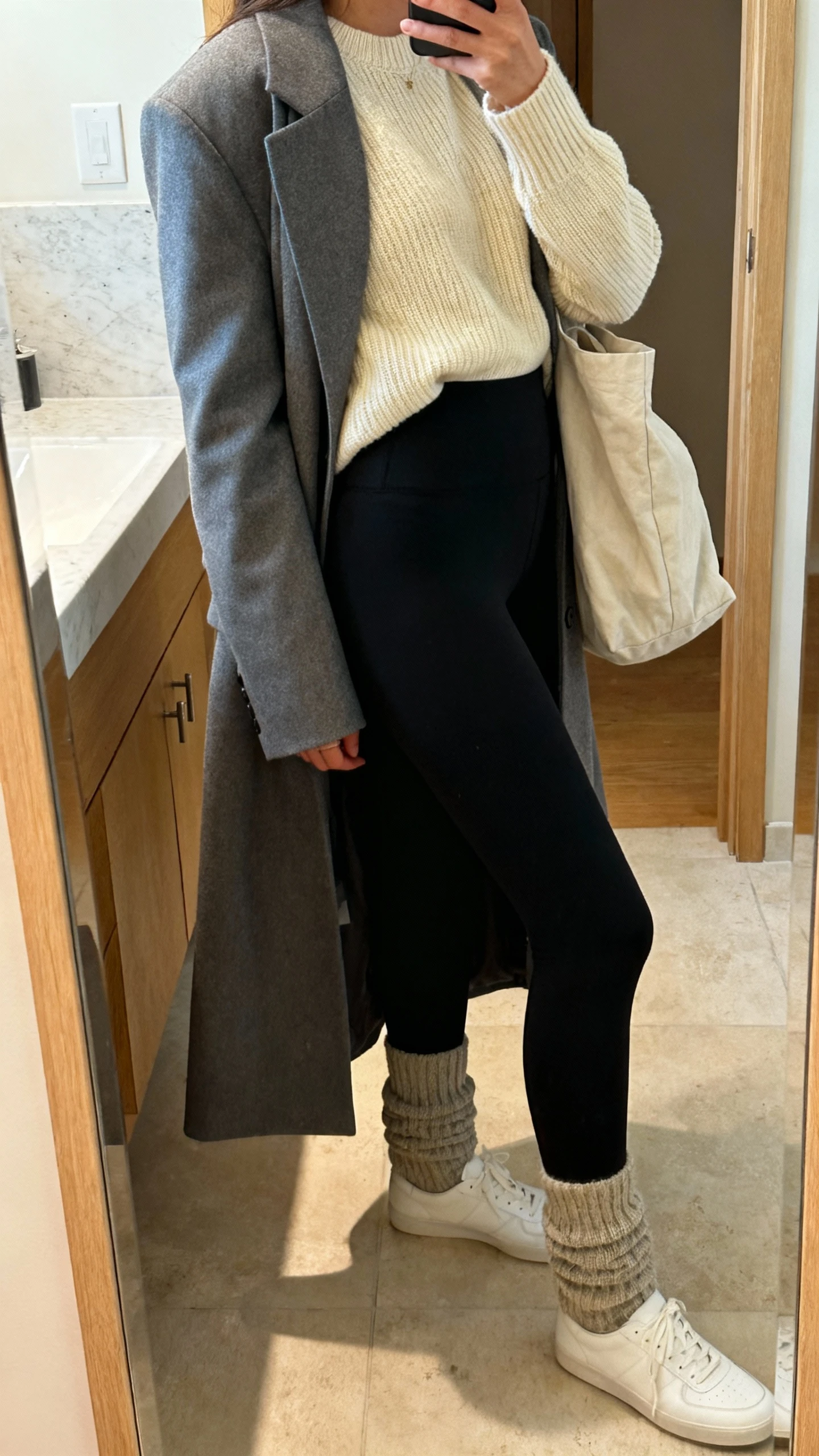 iPhone side-angle mirror selfie of a woman wearing a soft cream knit sweater, black high-waisted leggings, long gray blazer, chunky socks, minimalist white trainers, and a slouchy tote, face not visible, cropped at shoulders, bathroom mirror, warm indoor lighting, iPhone photo quality.