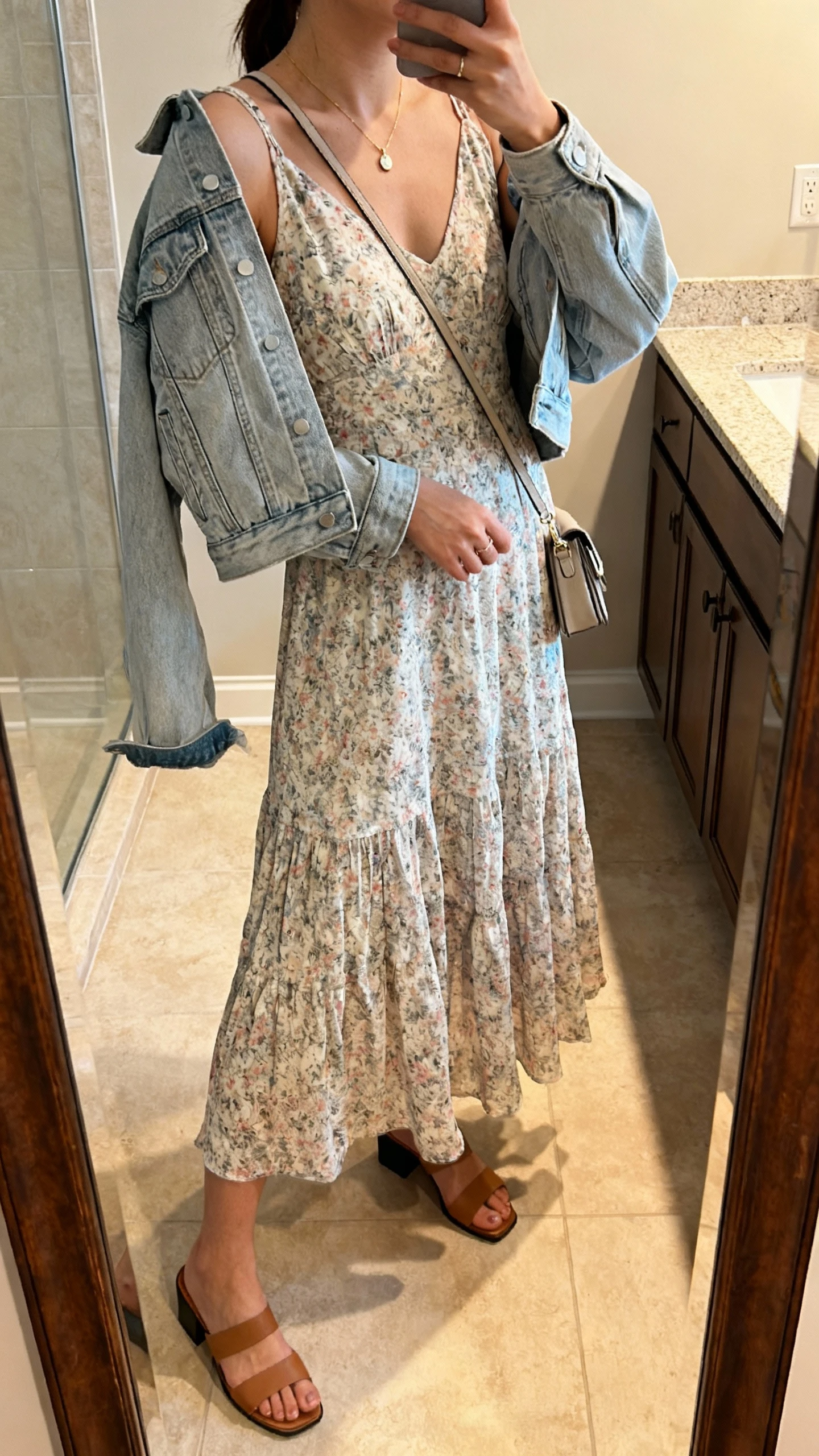 iPhone side-angle mirror selfie of a woman wearing a flowy floral midi dress with a light-wash denim jacket draped over shoulders, dainty pendant necklace, low block-heel sandals, small crossbody bag, face not visible, bathroom mirror, natural indoor lighting, iPhone photo quality