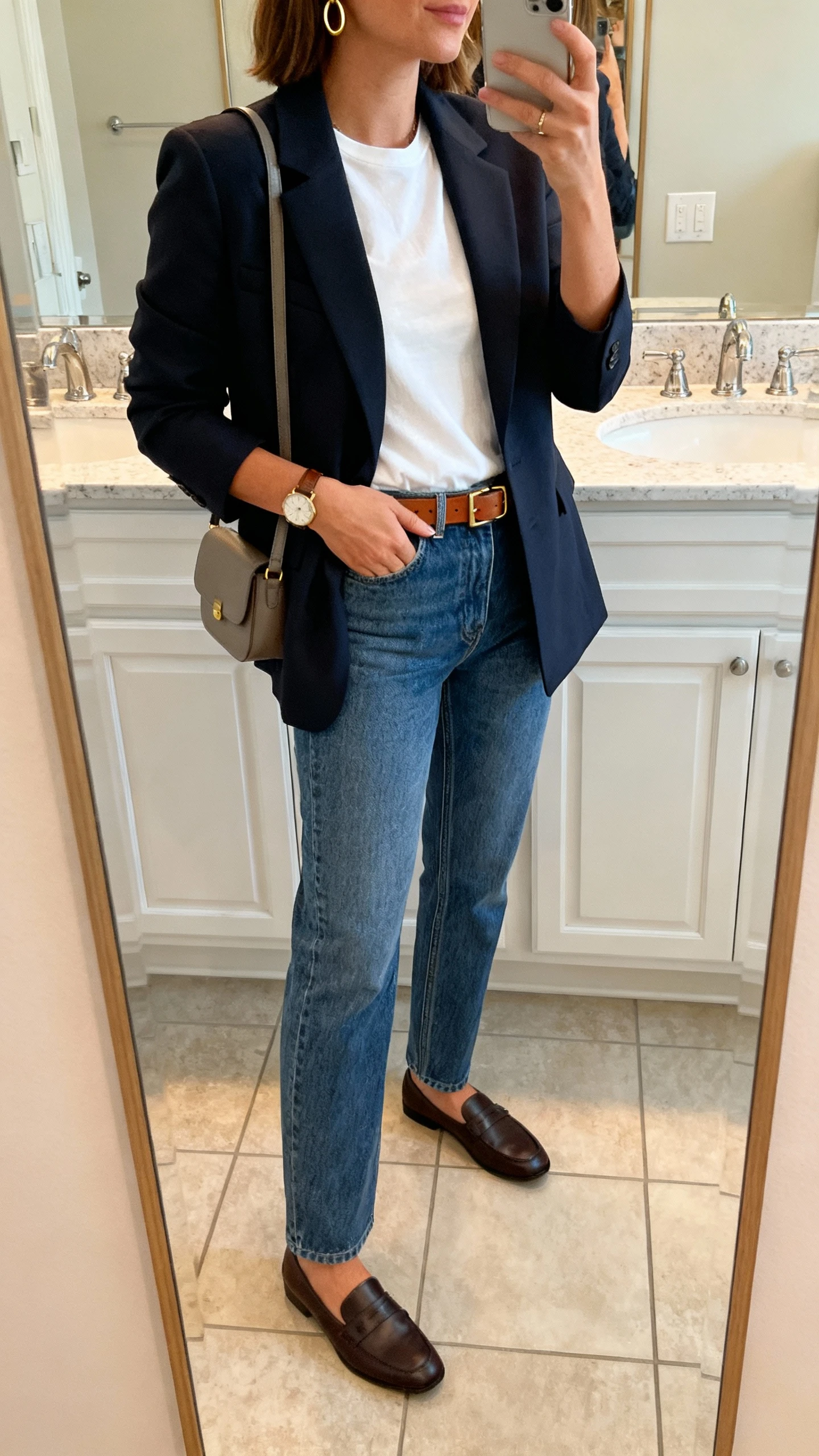 iPhone side-angle mirror selfie of a woman wearing straight-leg jeans, structured blazer, simple white tee, loafers, slim leather belt, small shoulder bag, minimal gold hoops and watch, face not visible, bathroom mirror, soft indoor lighting, iPhone photo quality.