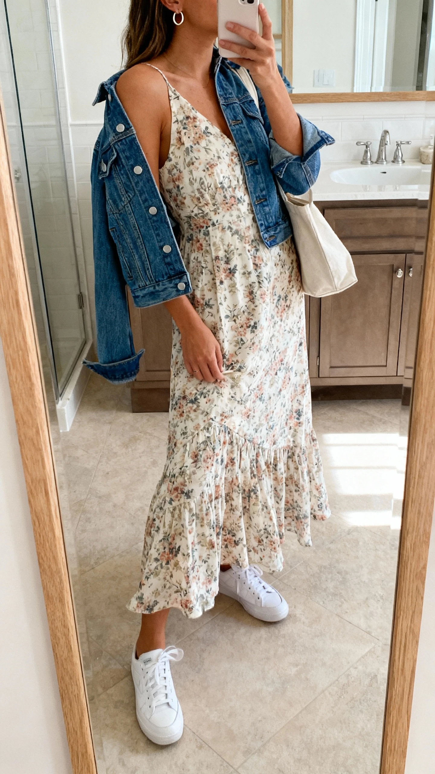 iPhone side-angle mirror selfie of a woman wearing a flowy floral midi dress paired with clean white sneakers, a denim jacket draped over the shoulders, small hoop earrings, and a simple tote bag, face not visible, bathroom full-length mirror, natural indoor daylight, iPhone photo quality