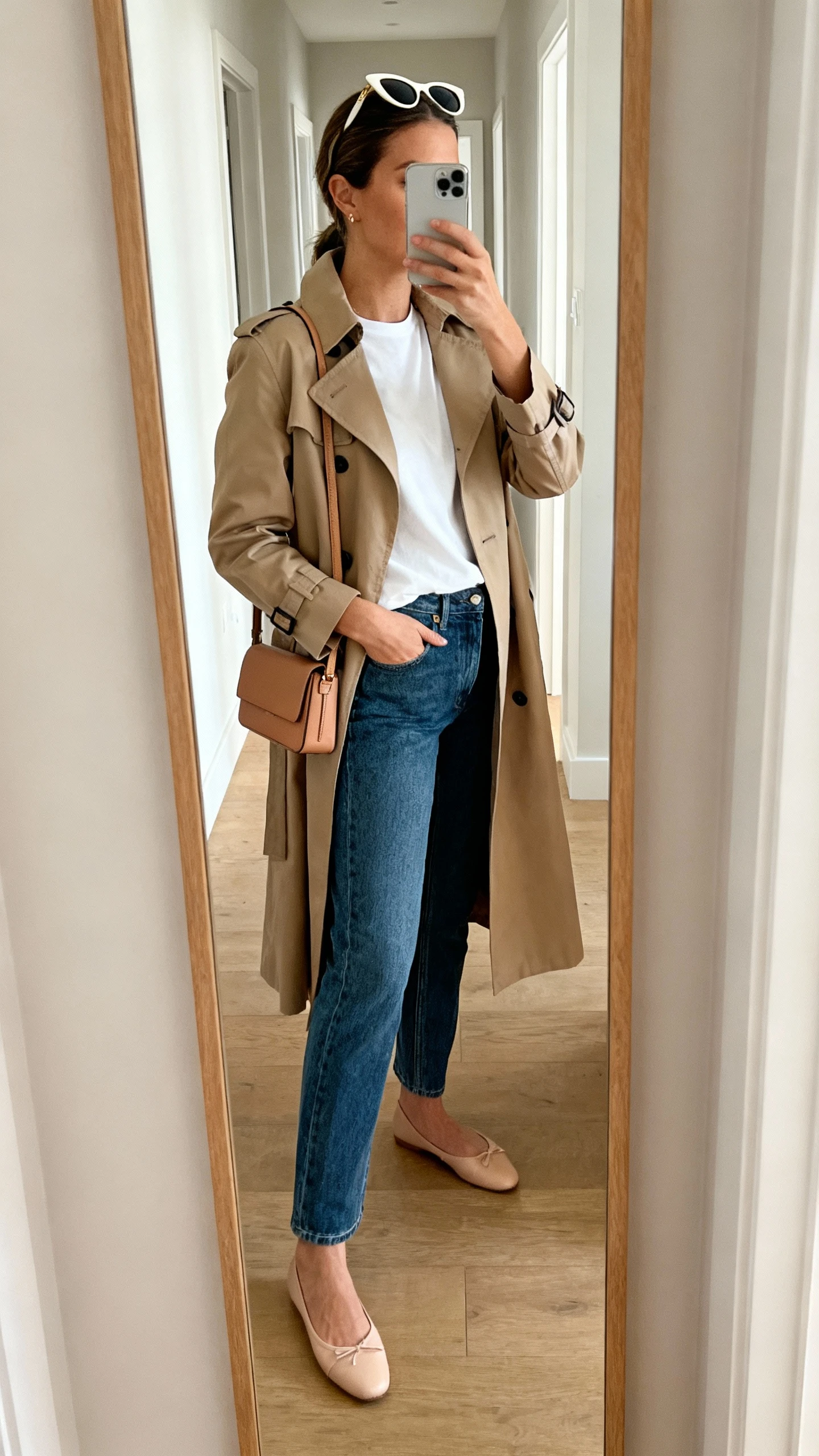 iPhone side-angle mirror selfie of a woman wearing a classic trench coat over a white tee with straight-leg jeans, ballet flats, slim leather shoulder bag, and cat-eye sunglasses pushed to the head, face not visible, cropped at shoulders, hallway mirror, natural daylight, iPhone photo quality.