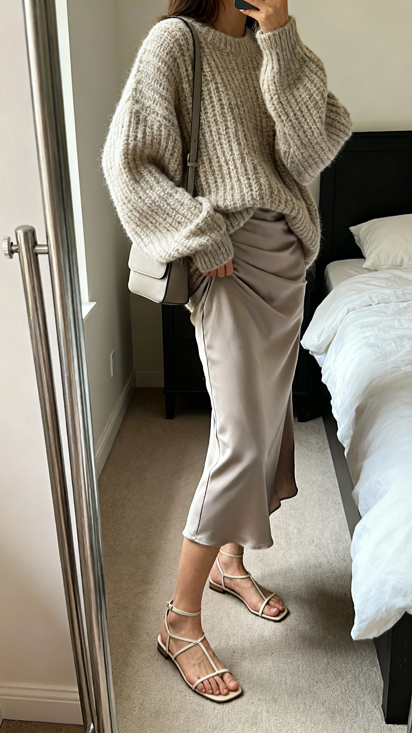 iPhone side-angle mirror selfie of a woman wearing a bias-cut slip dress layered under an oversized chunky knit sweater, paired with slim strap sandals and a simple shoulder bag, face not visible, cropped at shoulders, full-length mirror in bedroom, soft indoor lighting, iPhone photo quality.