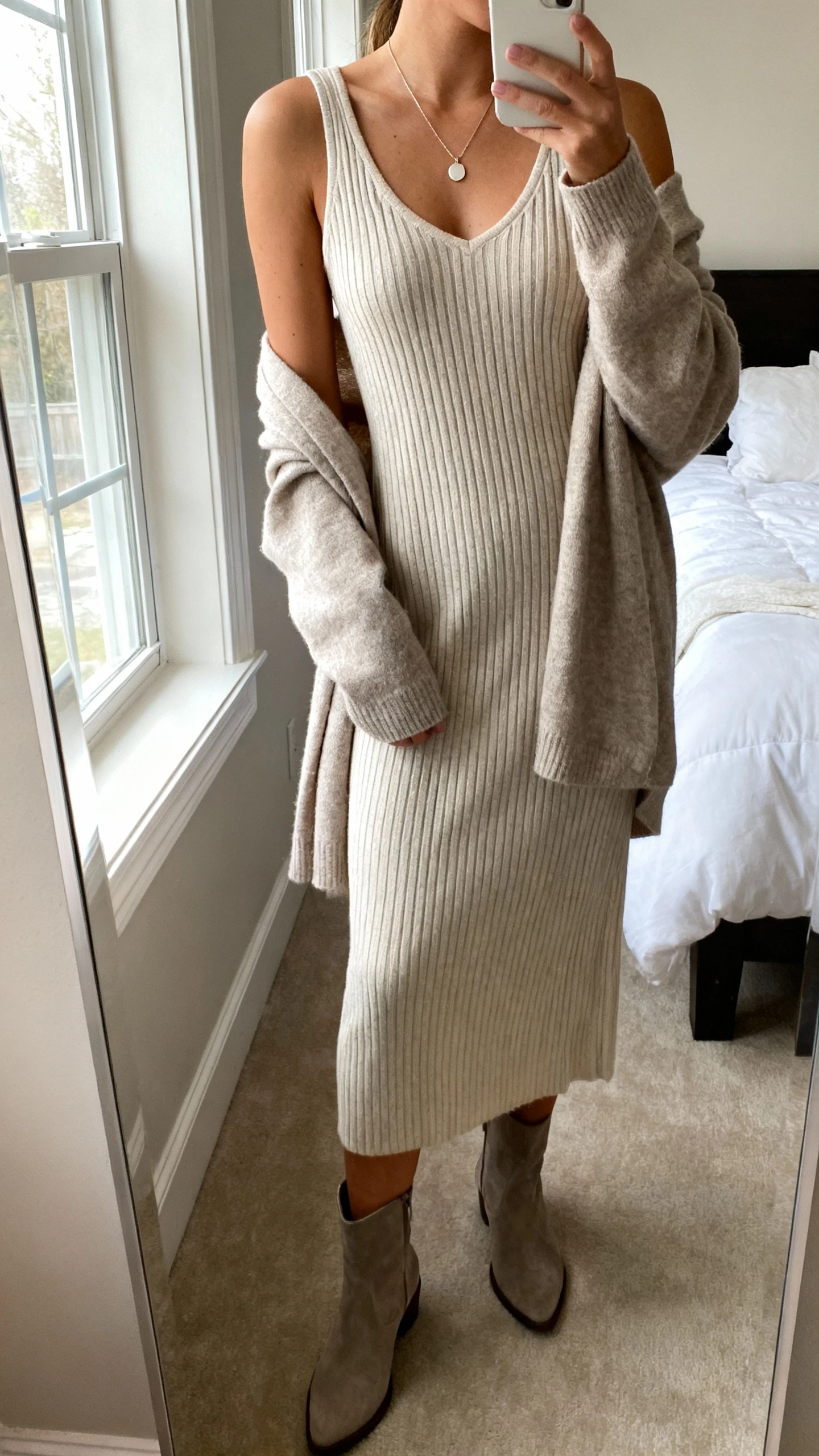 iPhone side-angle mirror selfie of a woman wearing a ribbed knit midi dress with a lightweight cardigan draped over shoulders, ankle boots, and a delicate pendant necklace, face not visible, cropped at shoulders, bedroom mirror, natural window light, iPhone photo quality.