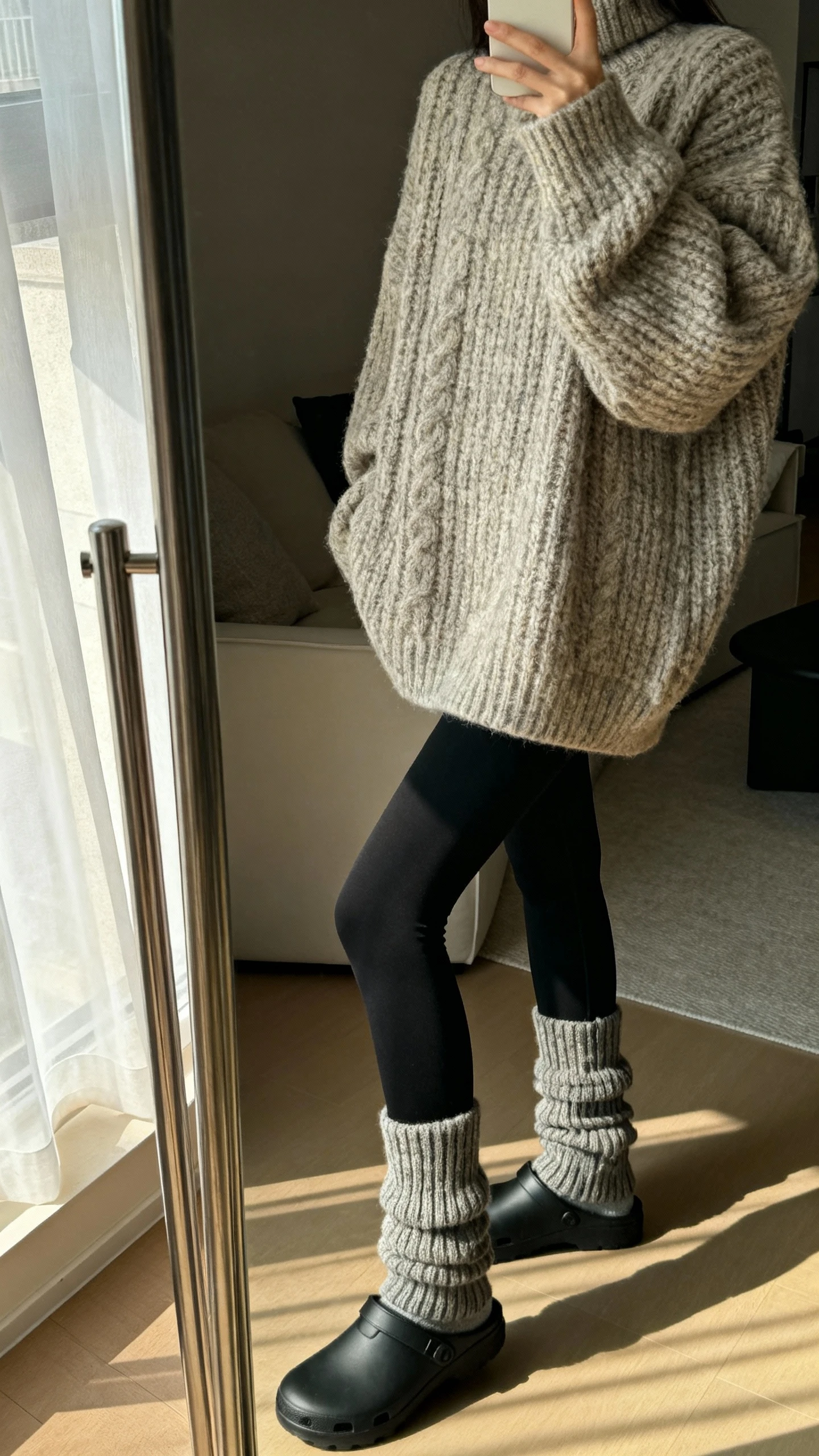iPhone side-angle mirror selfie of a woman wearing an oversized chunky sweater with black leggings, chunky socks and slip-on clogs, face not visible, cropped at shoulders, living room mirror, soft afternoon window light, iPhone photo quality.