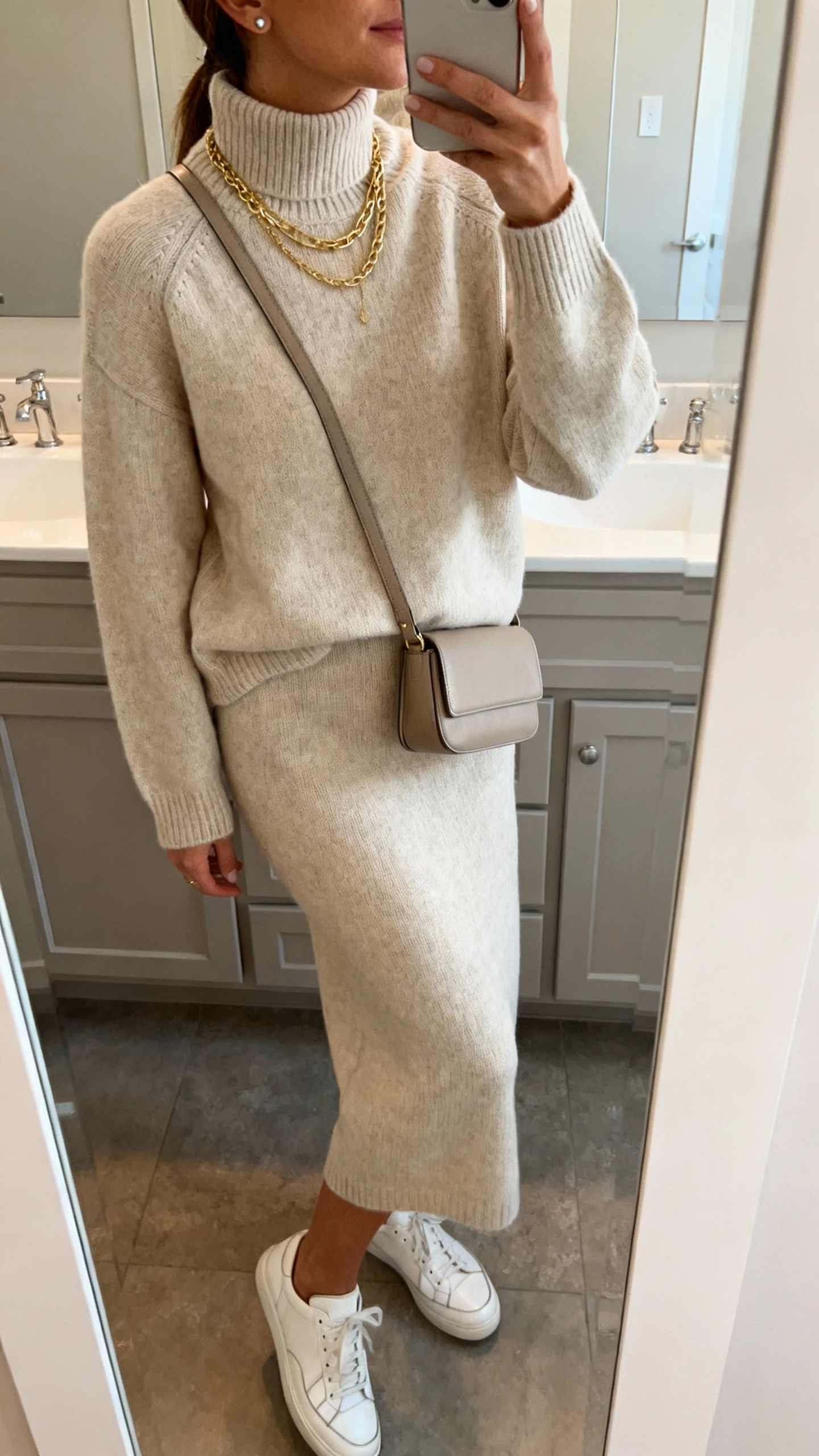 iPhone side-angle mirror selfie of a woman wearing a monochrome knit set: light beige merino mock-neck sweater with matching knit midi skirt, sleek white leather sneakers, a structured crossbody, layered gold necklaces and simple studs, face not visible, bathroom mirror, soft indoor lighting, iPhone photo quality.