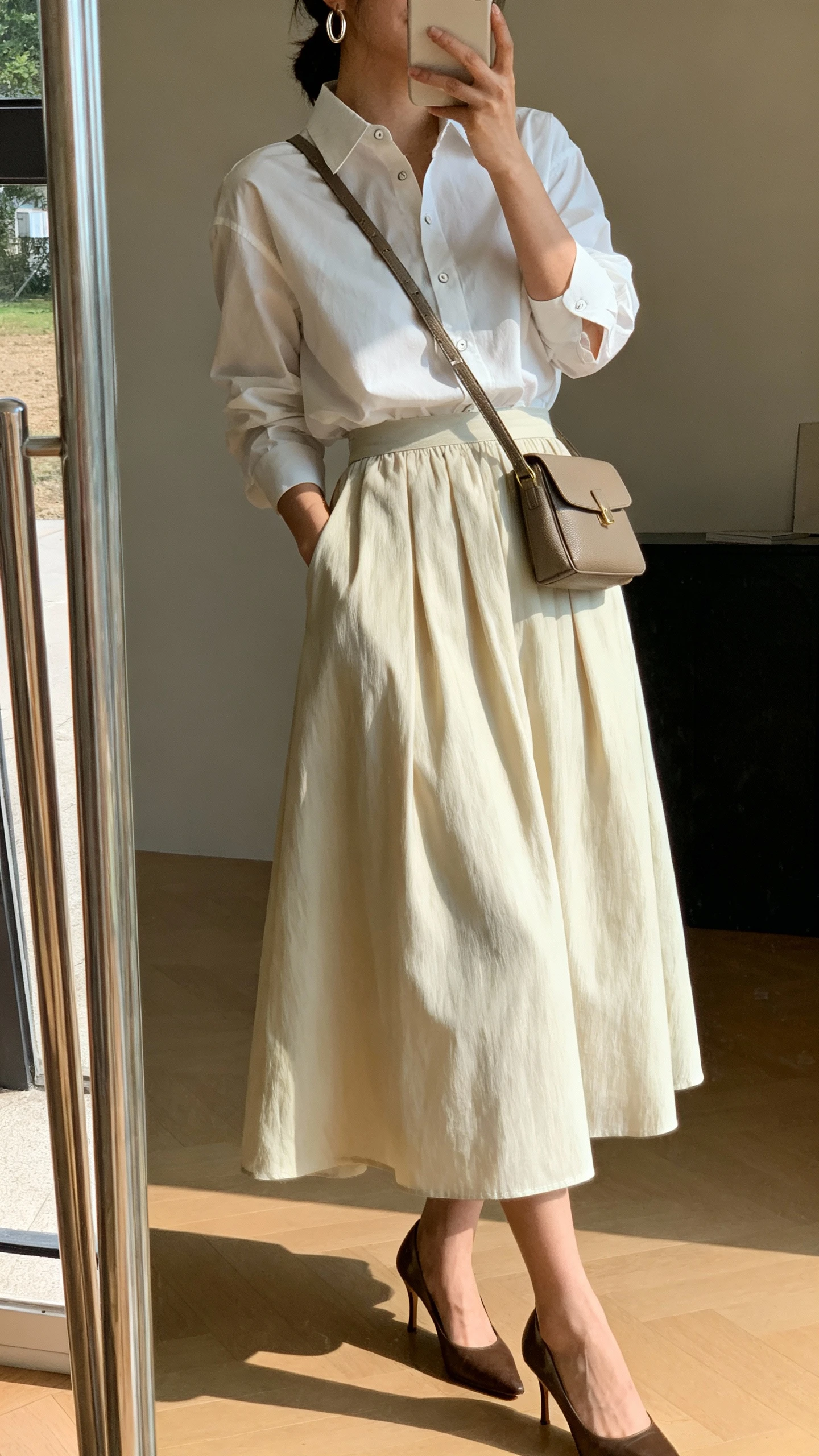 iPhone side-angle mirror selfie of a woman wearing a crisp button-up shirt tucked into a flowy midi skirt, classic pumps, structured crossbody, simple hoop earrings, face not visible, cropped at shoulders, full-length mirror, indoor daylight, iPhone photo quality.