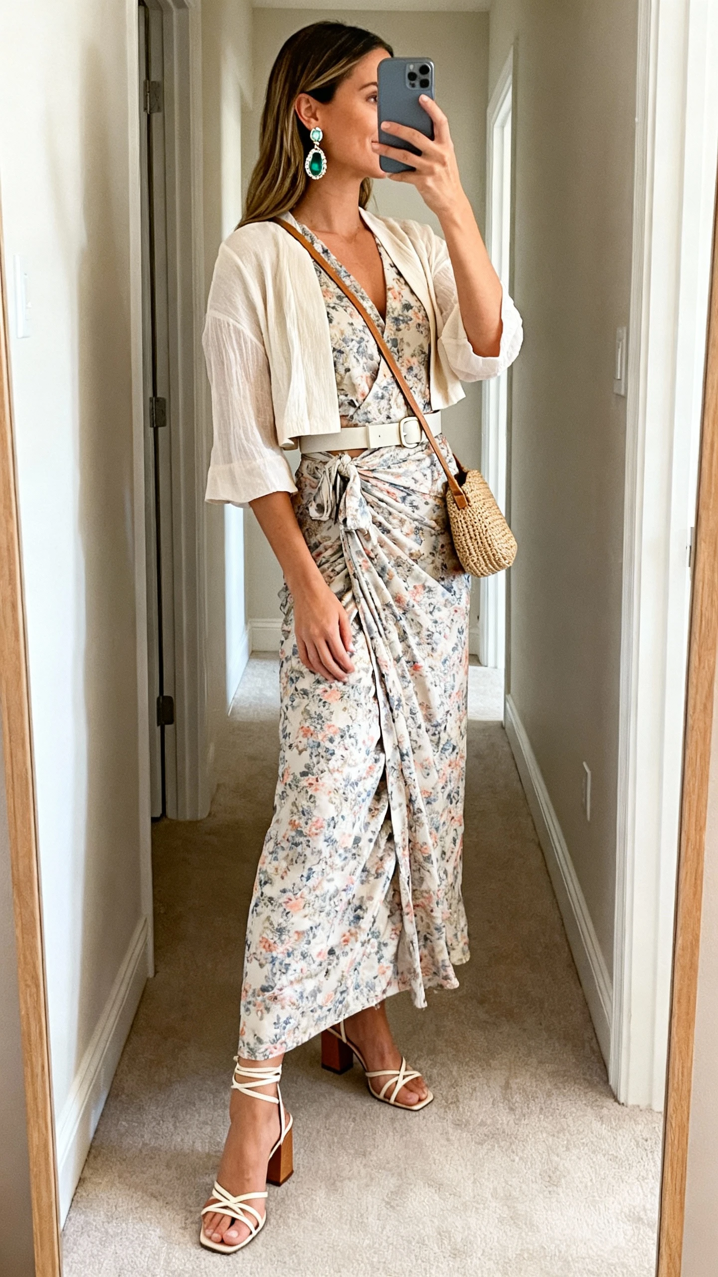 iPhone side-angle mirror selfie of a woman wearing a printed wrap maxi dress with a lightweight cropped cardigan, strappy block-heel sandals, statement gemstone drop earrings, a woven crossbody bag, and a soft belt at the waist; face not visible, full-length mirror in hallway, natural daylight, iPhone photo quality.
