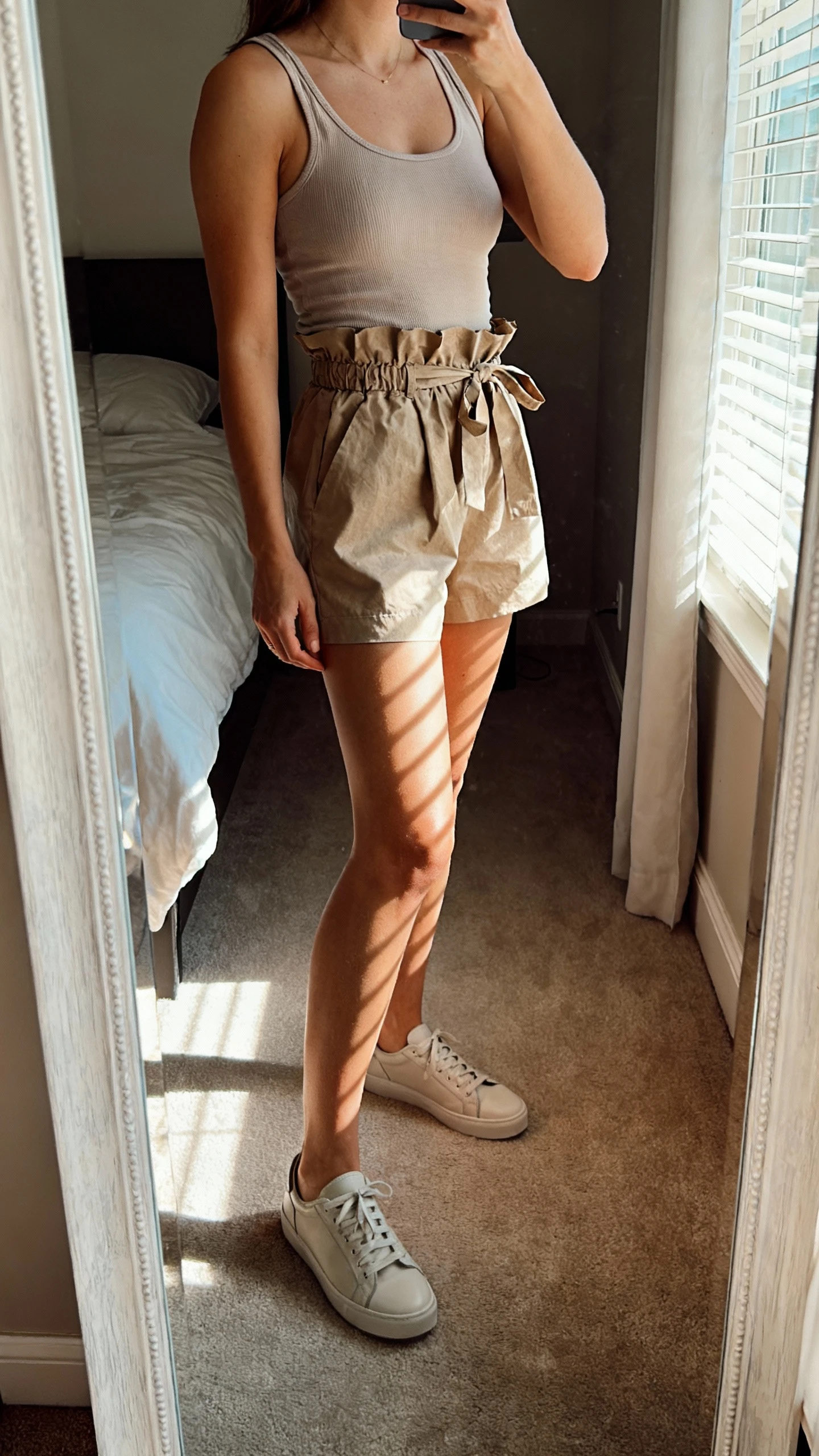 iPhone side-angle mirror selfie of a woman wearing a fitted tank, paperbag-waist shorts tied at the waist, and sleek minimalist sneakers, face not visible, bedroom mirror, afternoon window light, iPhone photo quality