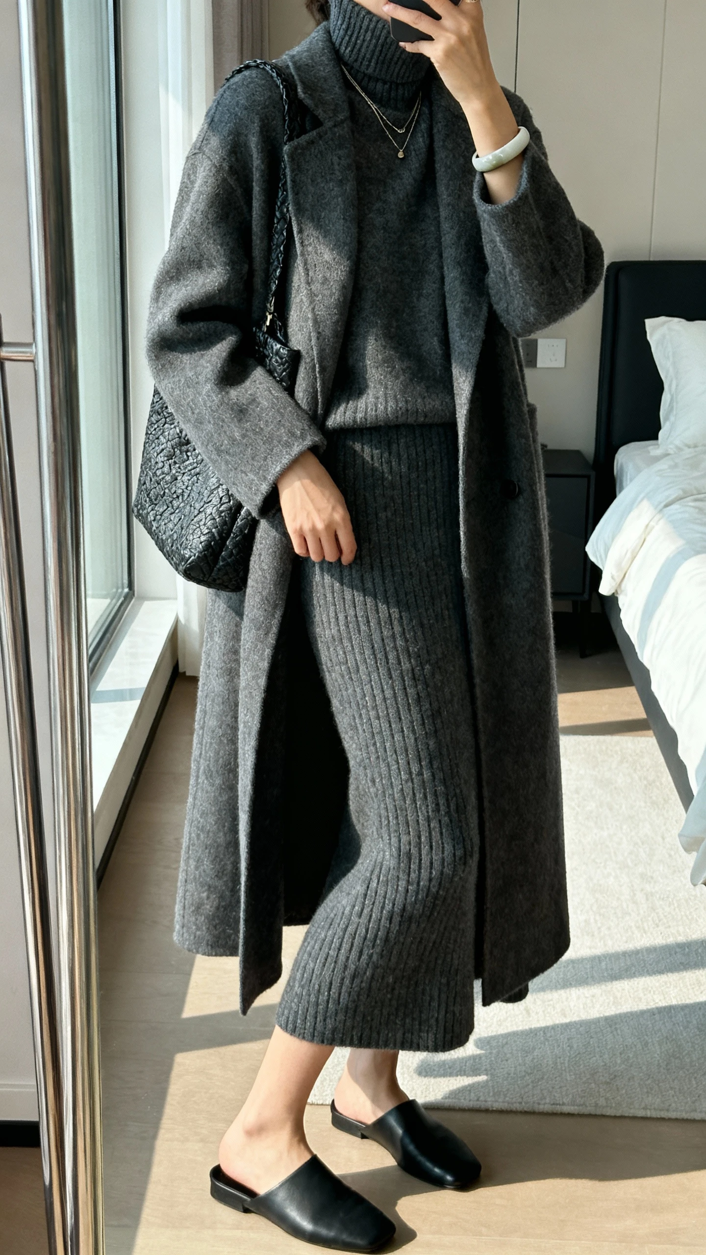 iPhone side-angle mirror selfie of a woman wearing a monochrome knit set: charcoal turtleneck sweater with matching knit midi skirt, minimalist black mules, longline wool coat, textured shoulder bag, subtle layered necklaces and a cuff bracelet, face not visible, bedroom mirror, gentle window light, iPhone photo quality.